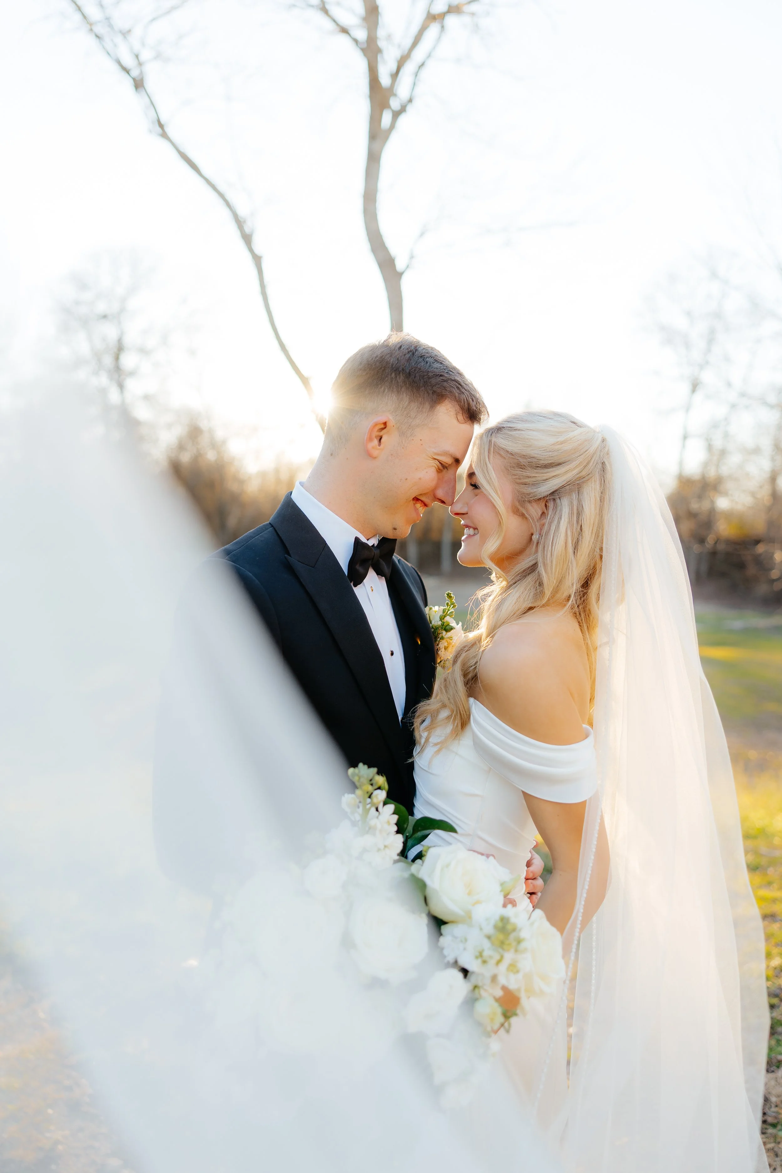 A bride and groom in wedding attire standing close together outdoors, touching foreheads with smiles, surrounded by trees and sunlight in the background.