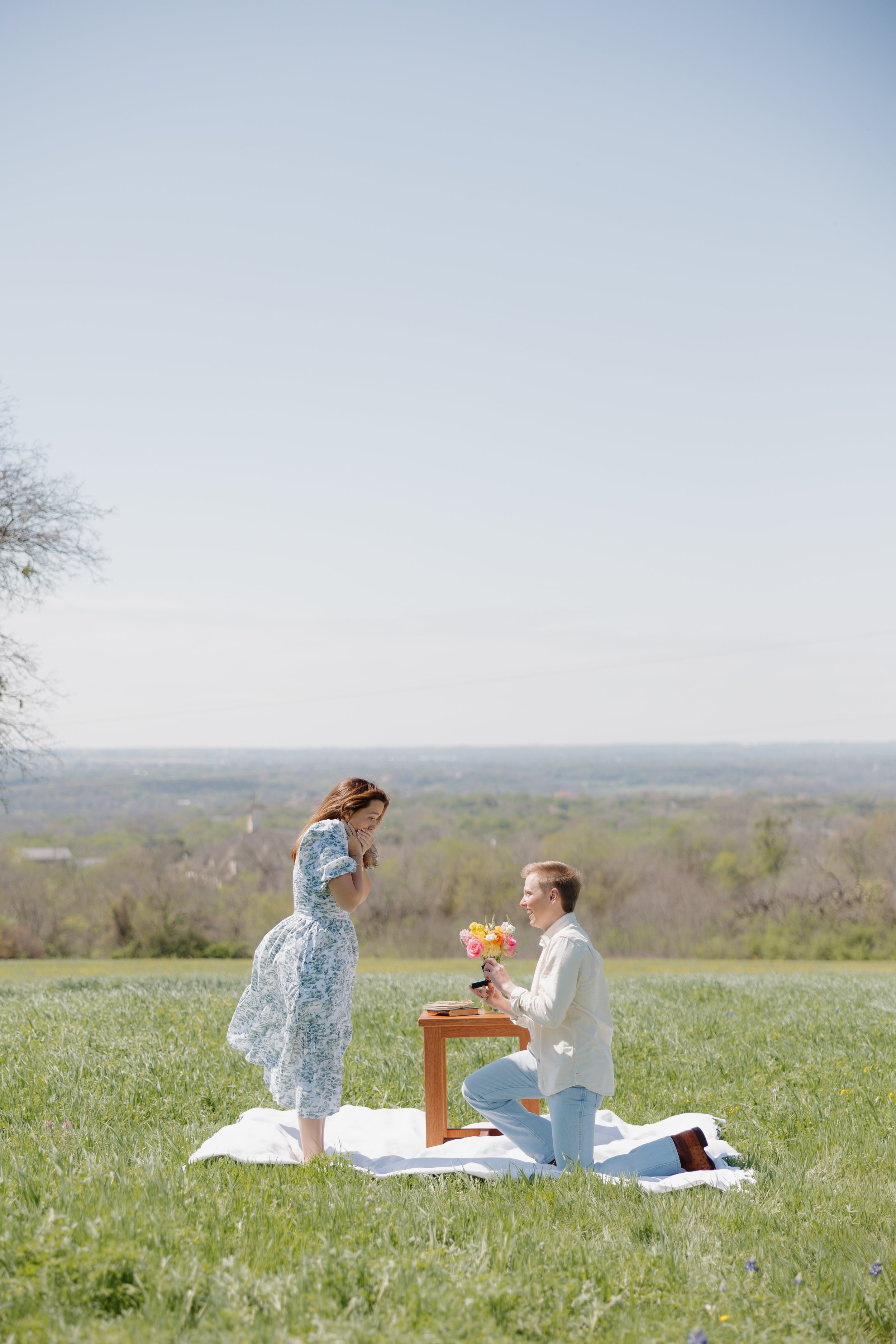 A man on one knee proposing marriage to a woman in an open field, with a woman holding her face in surprise. A small table with flowers and a ring box is set between them on a white blanket.