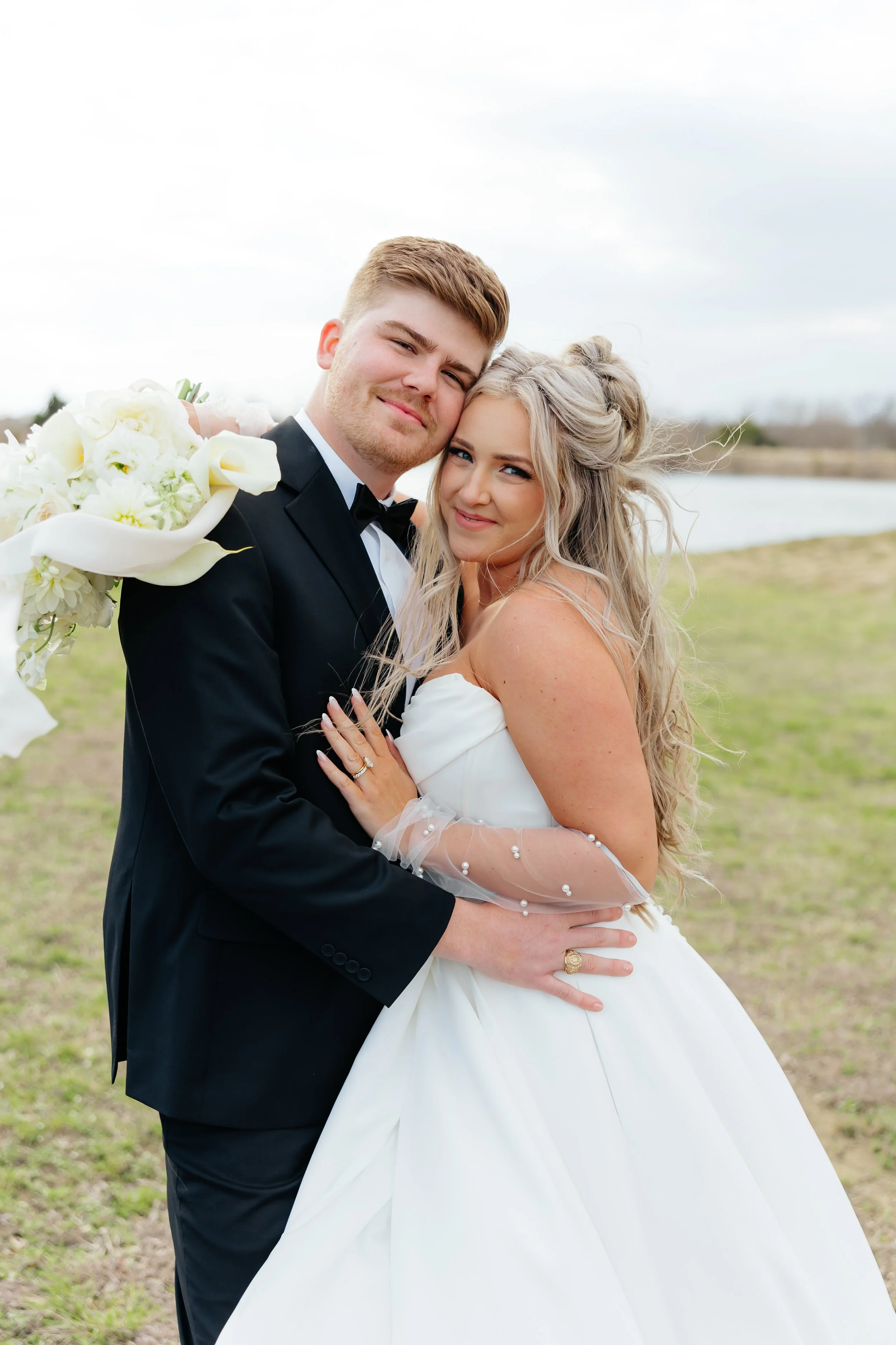 A newlywed couple standing outdoors near a body of water, dressed in wedding attire, smiling and embracing.