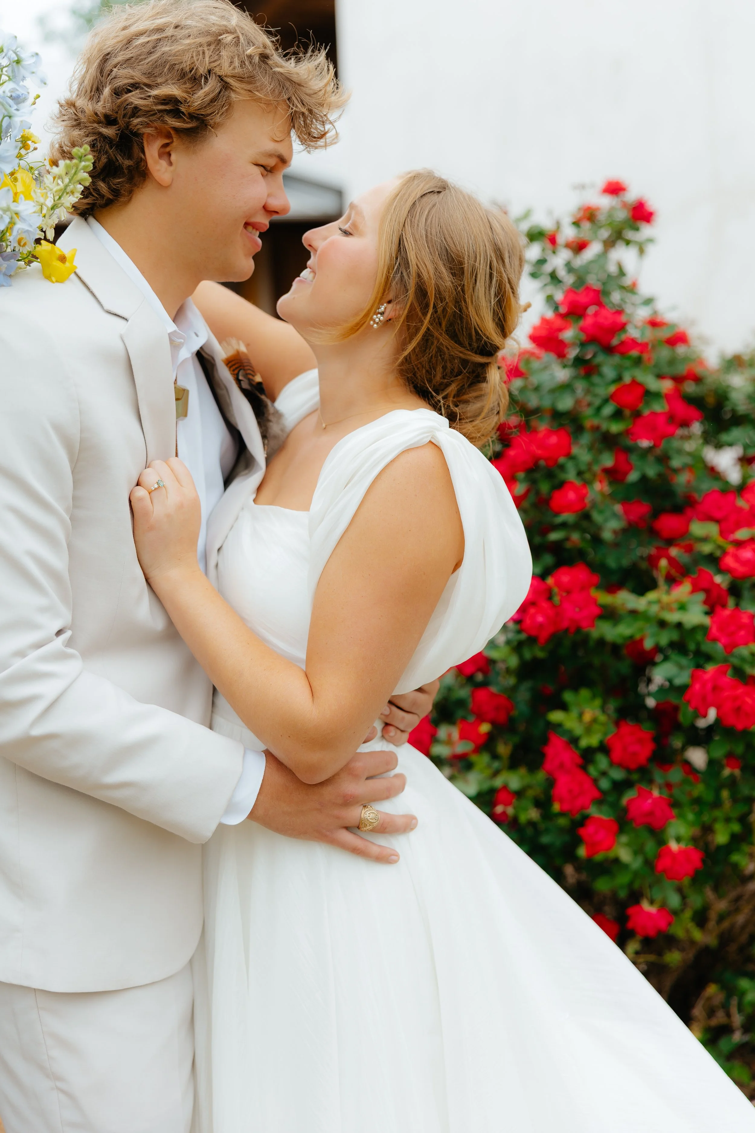 A happy bride and groom in wedding attire sharing a joyful moment outdoors, with blooming red flowers in the background.