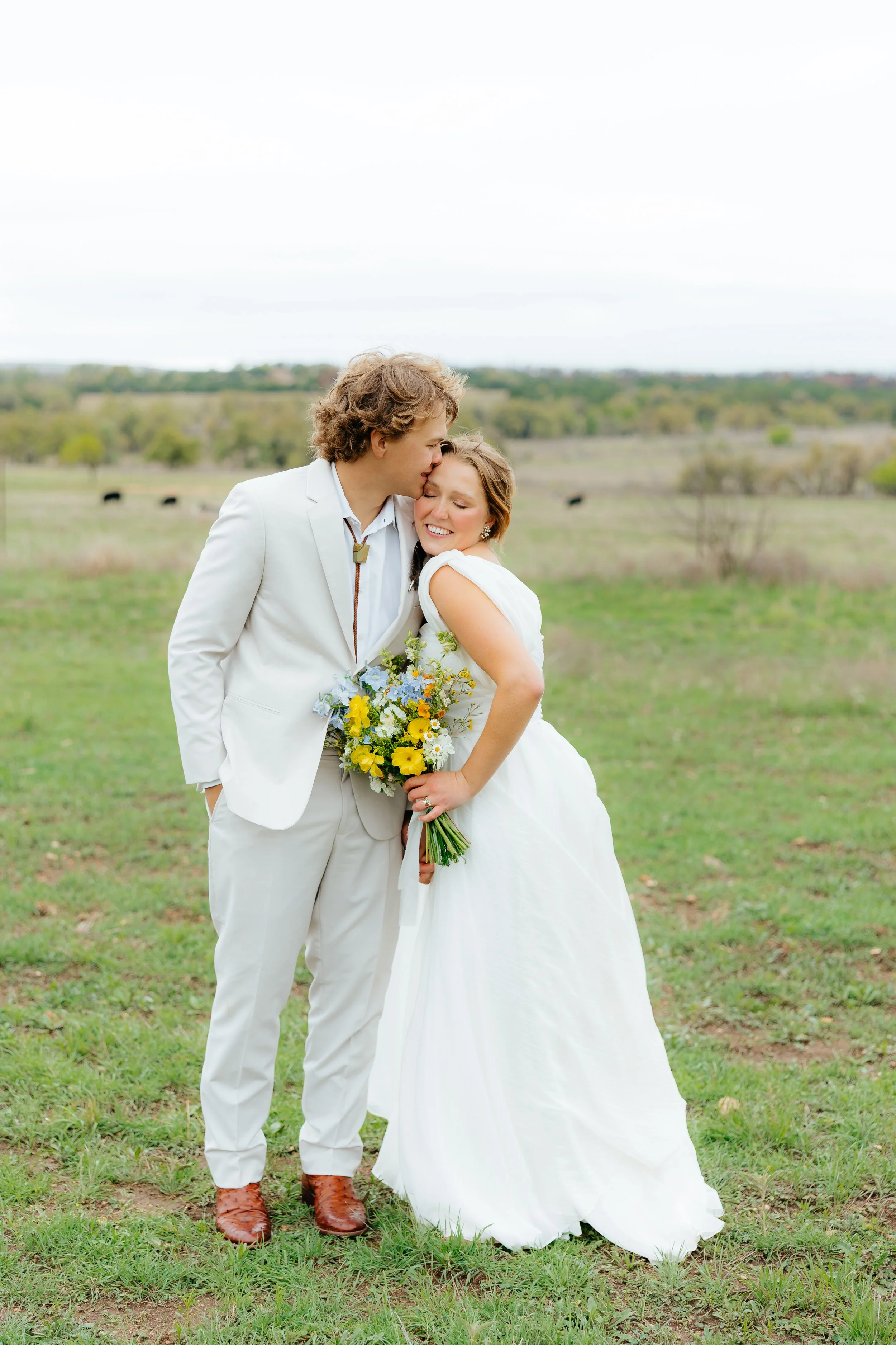 A newlywed couple stands outdoors in a grassy field, with the groom kissing the bride on the forehead and she smiling happily, holding a colorful bouquet of flowers.