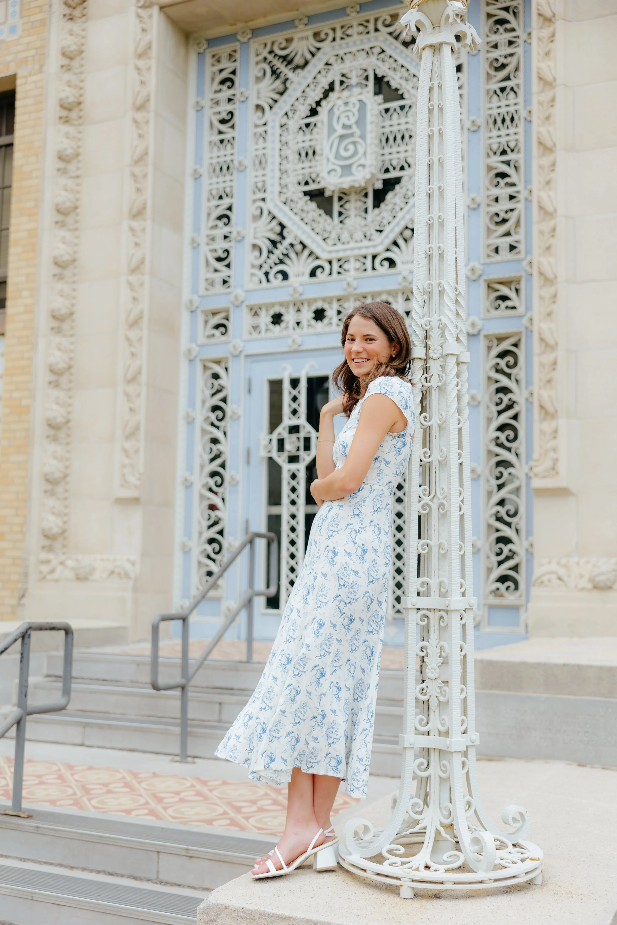 A young woman in a white and blue floral dress and white heels leaning against an ornate white metal structure outside a building with decorative stonework and a blue door.