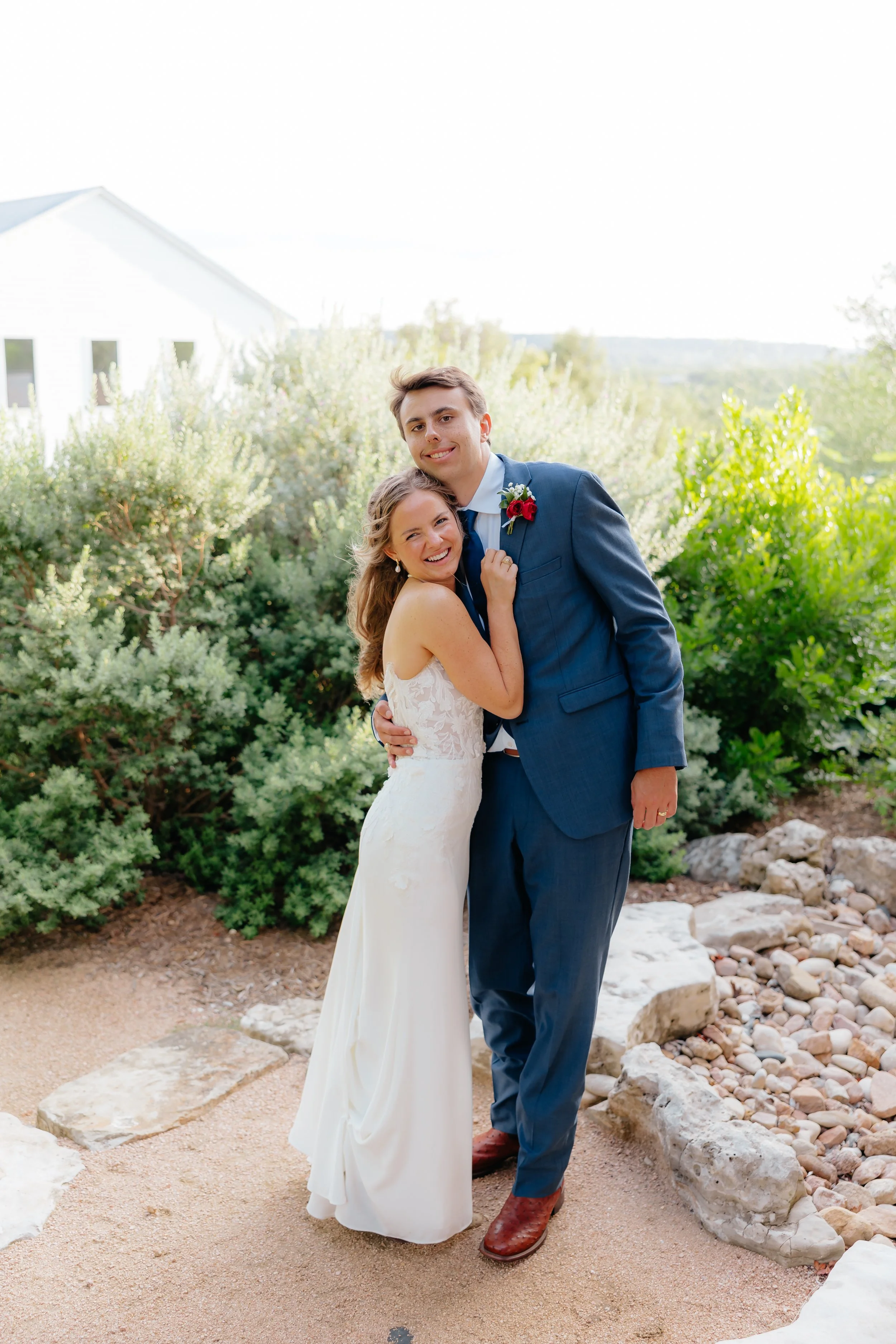 A bride and groom smiling and hugging each other outdoors during their wedding, with greenery and rocks in the background.