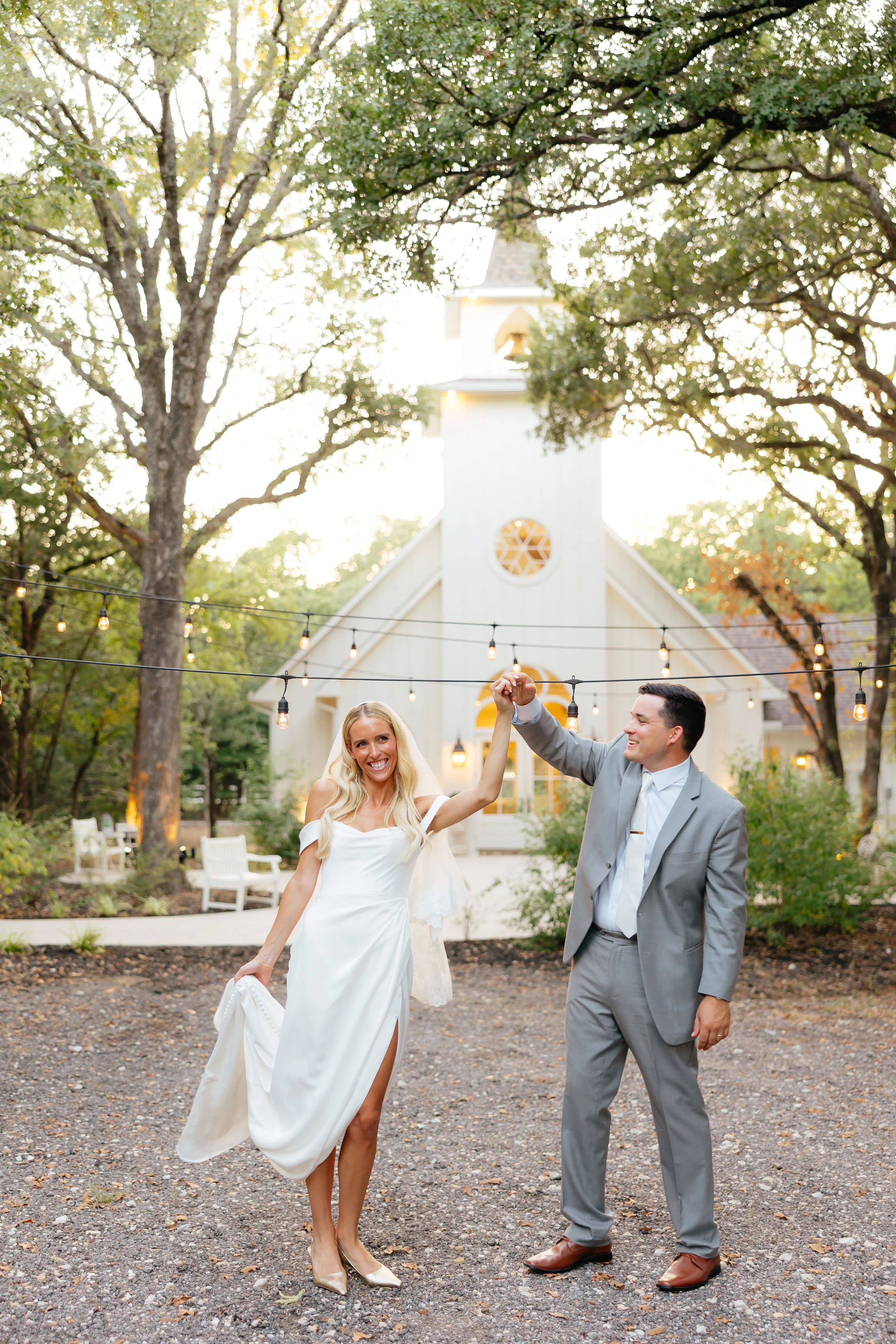 A bride and groom dancing outdoors in front of a church with string lights and trees.