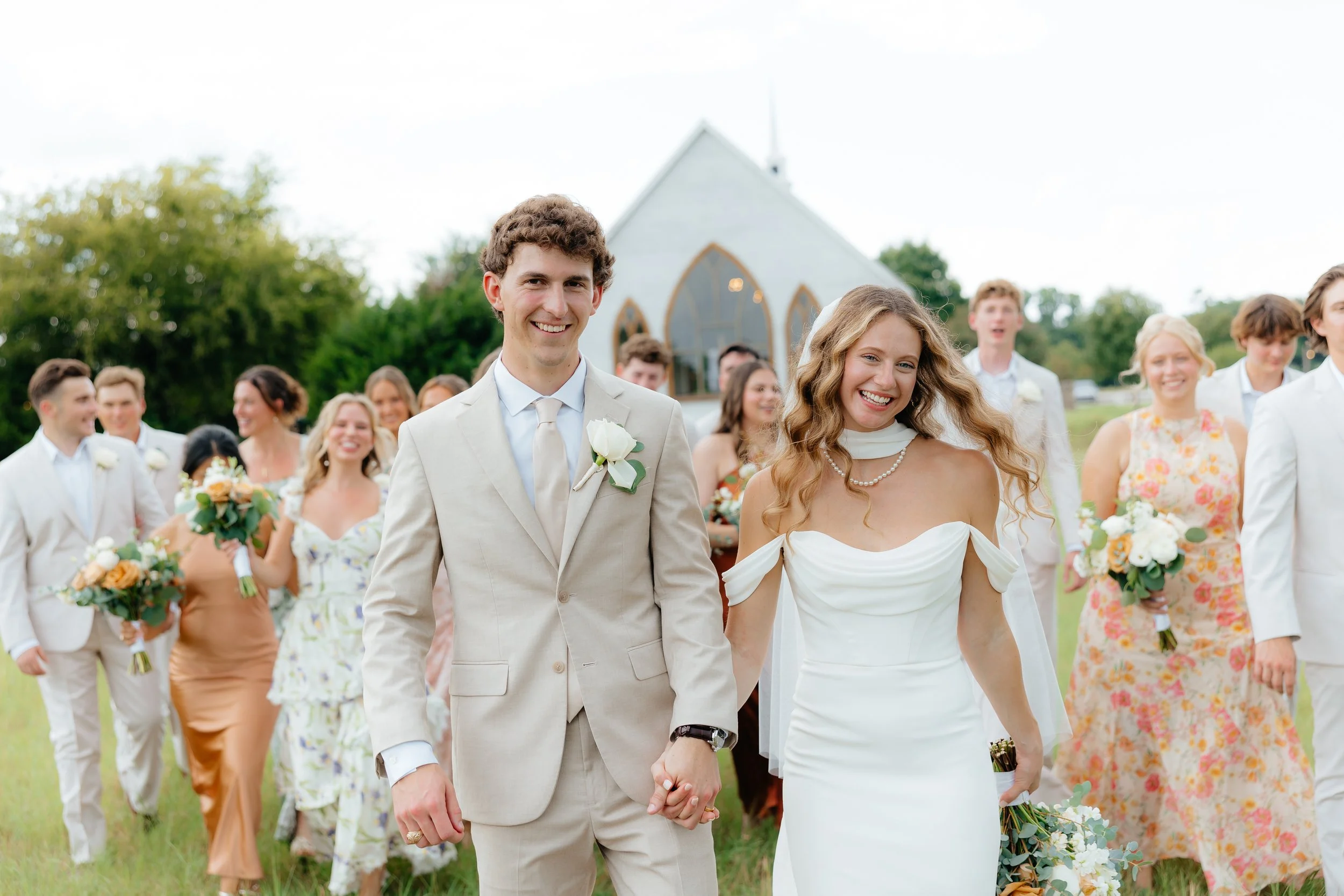 A bride and groom walking hand-in-hand outdoors with their wedding party, in front of a small church, during daytime.