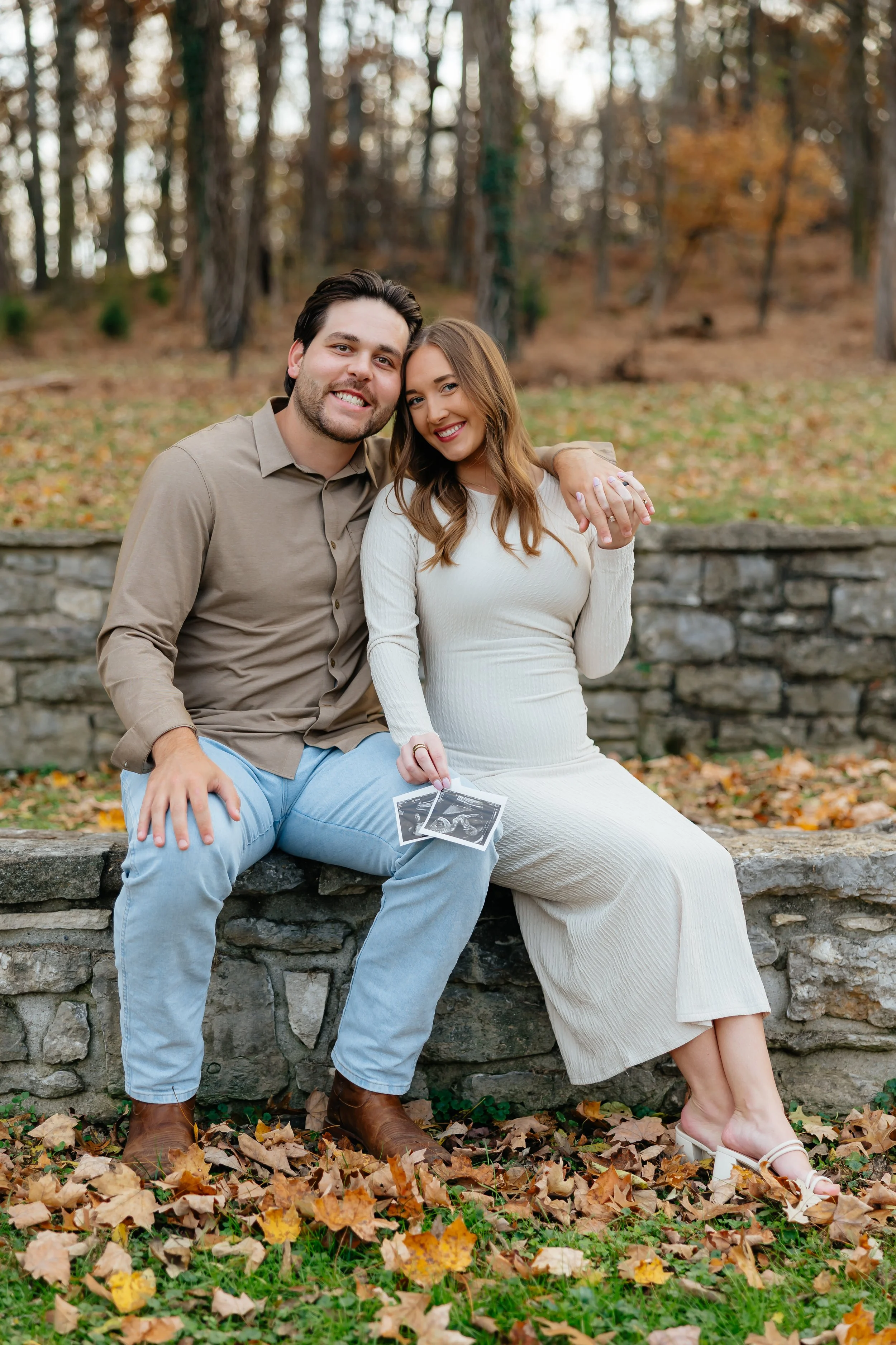 A couple sitting on a stone wall in a park during autumn, smiling, with the woman holding ultrasound photos and the man placing his arm around her shoulder.