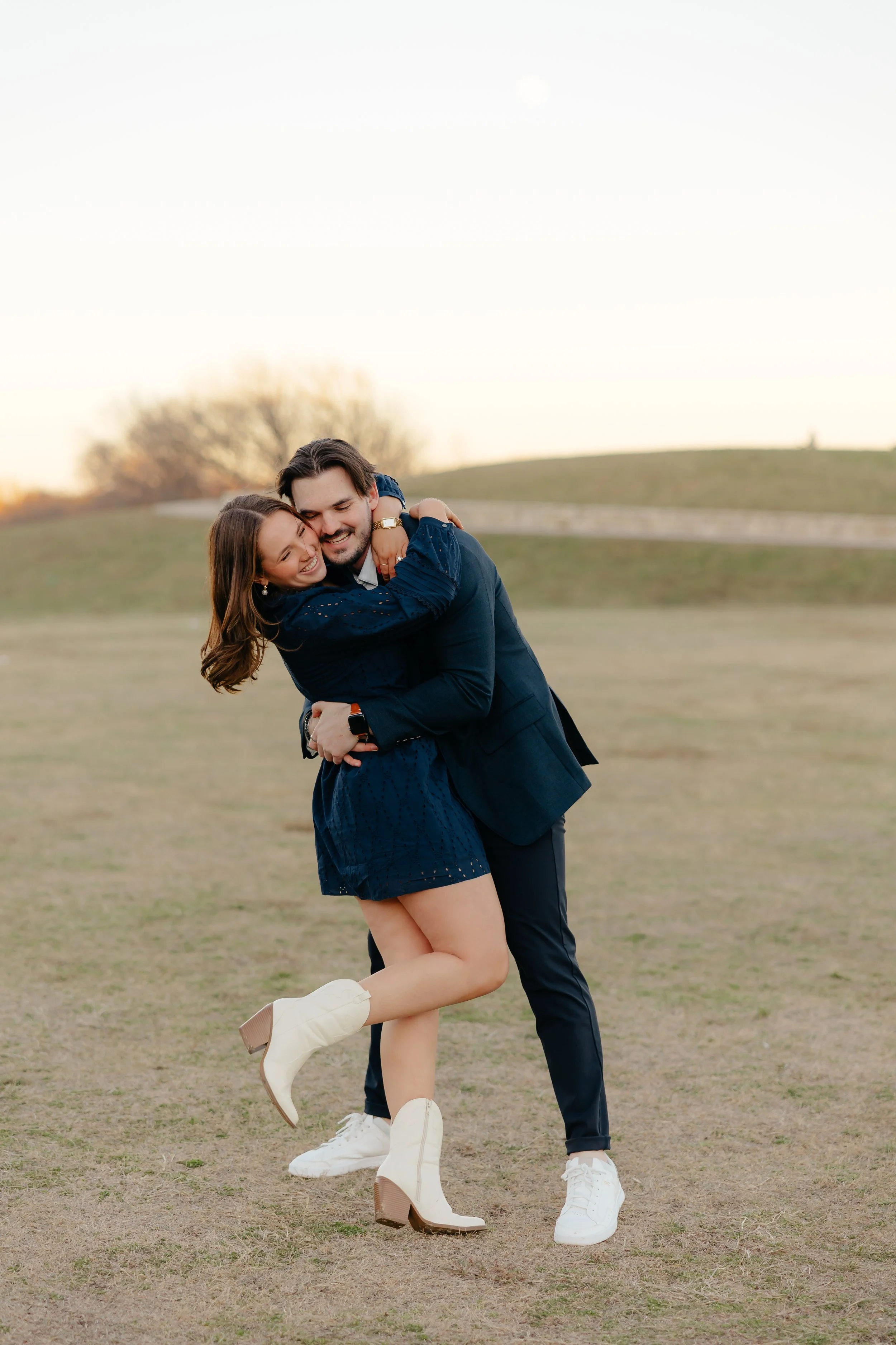 A happy couple hugging and smiling outdoors on a grassy field during sunset, the woman wearing a navy dress and white boots, the man in a dark suit with white sneakers.