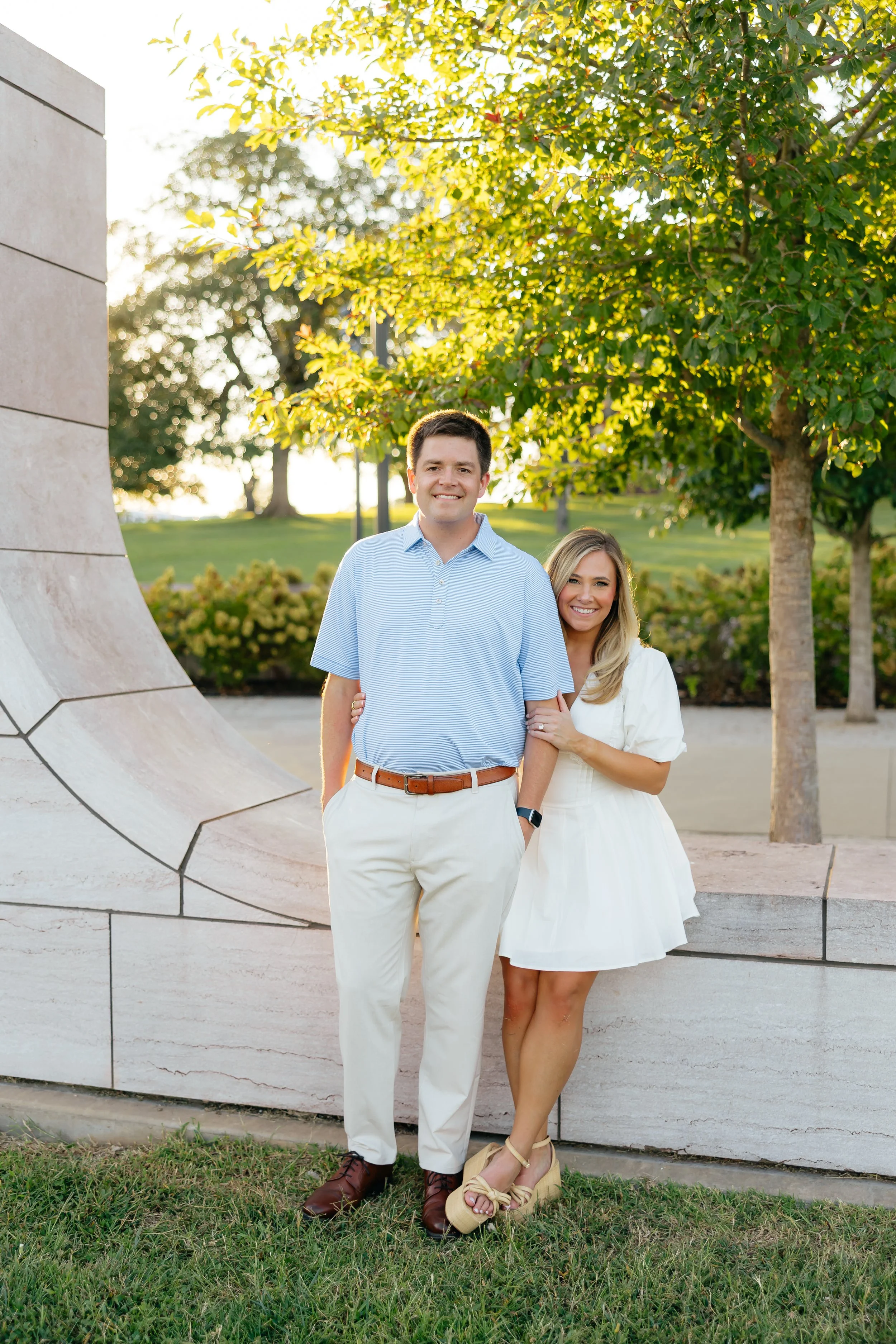 A smiling couple stands outdoors near a stone structure, with trees and greenery in the background, during sunset.