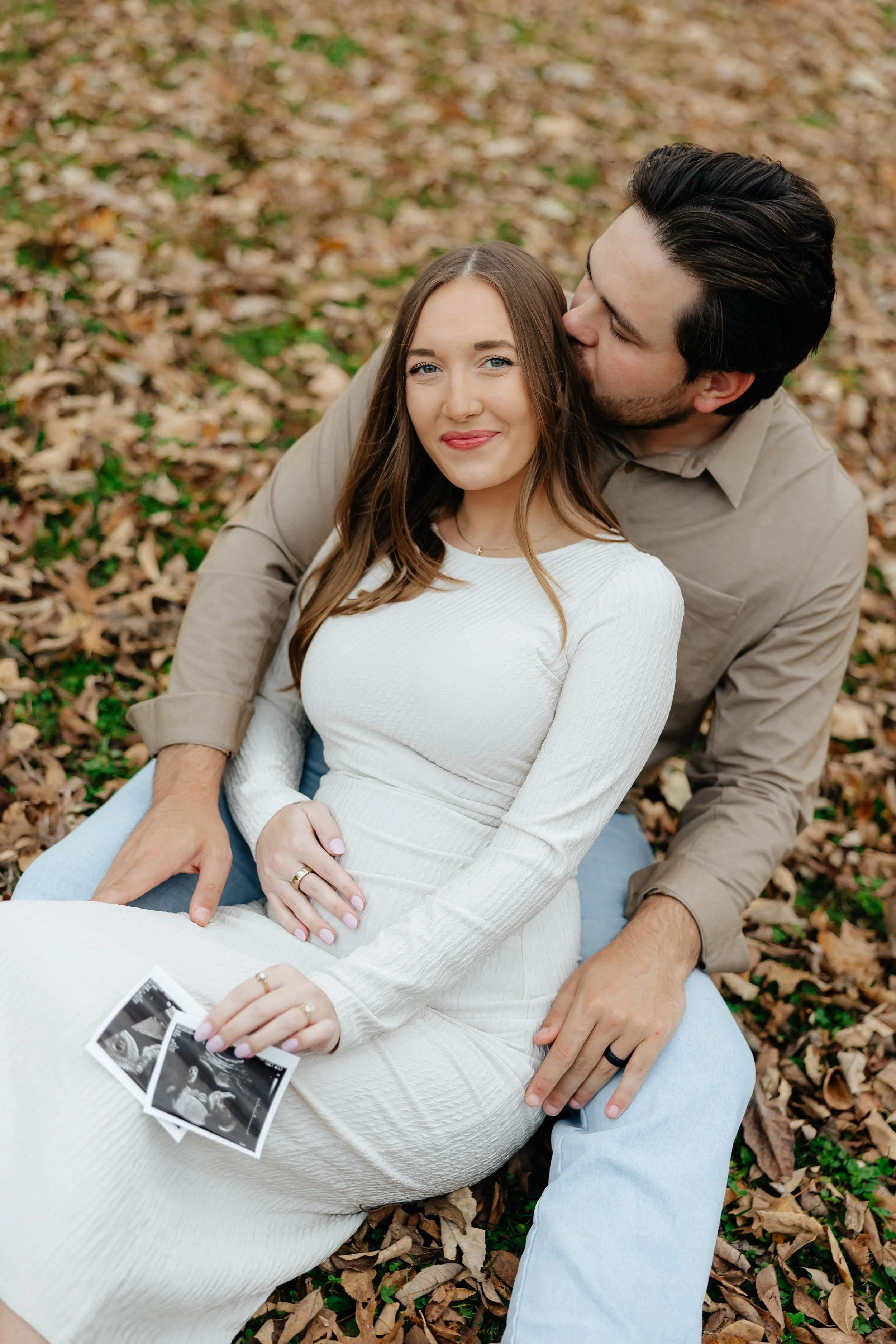 A young couple sitting on fallen leaves outdoors, holding ultrasound photos. The woman is smiling while the man kisses her on the side of the head.