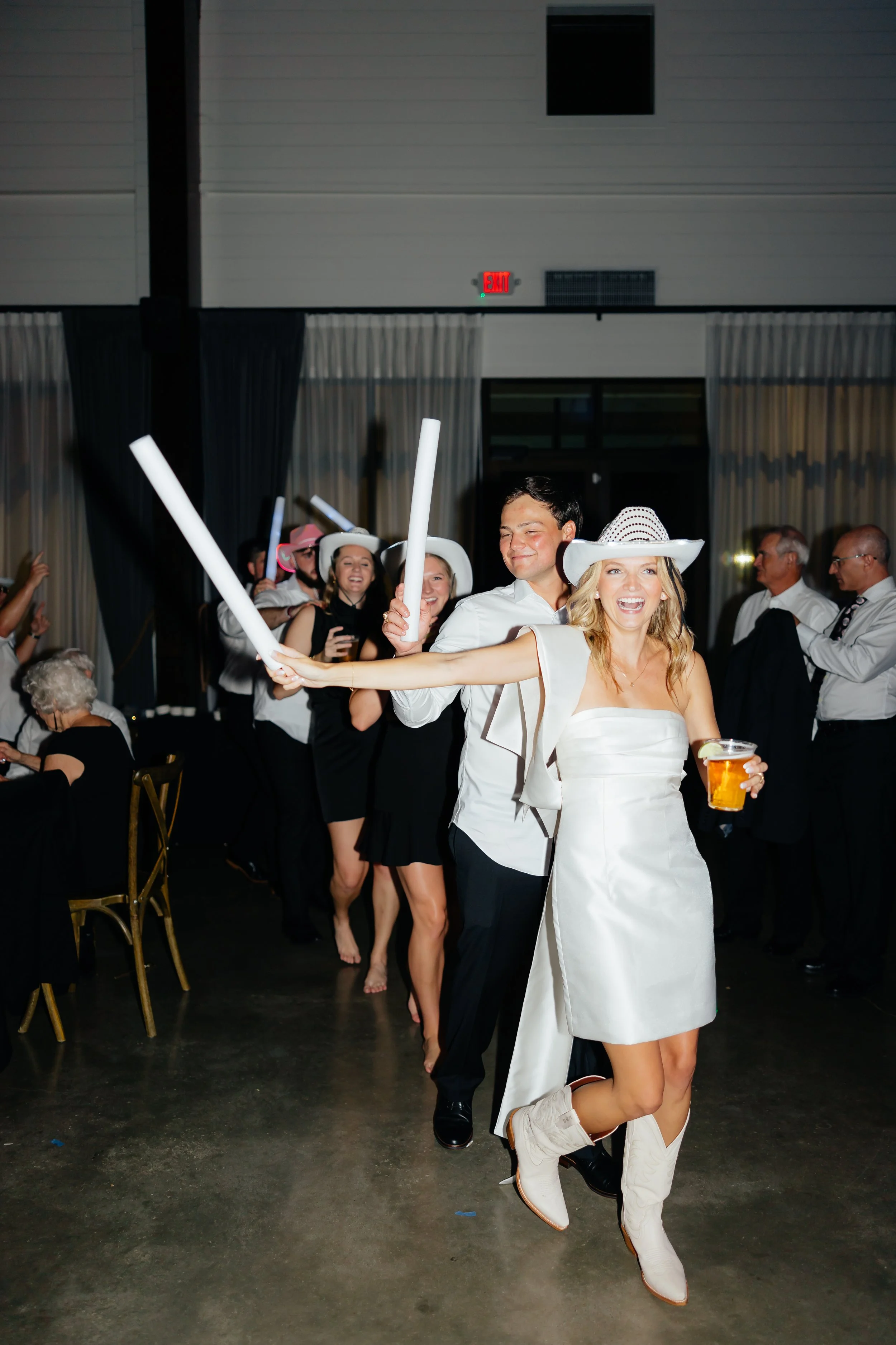 People dressed in Western-themed costumes including cowboy hats, holding foam sticks, and dancing at a party or celebration.