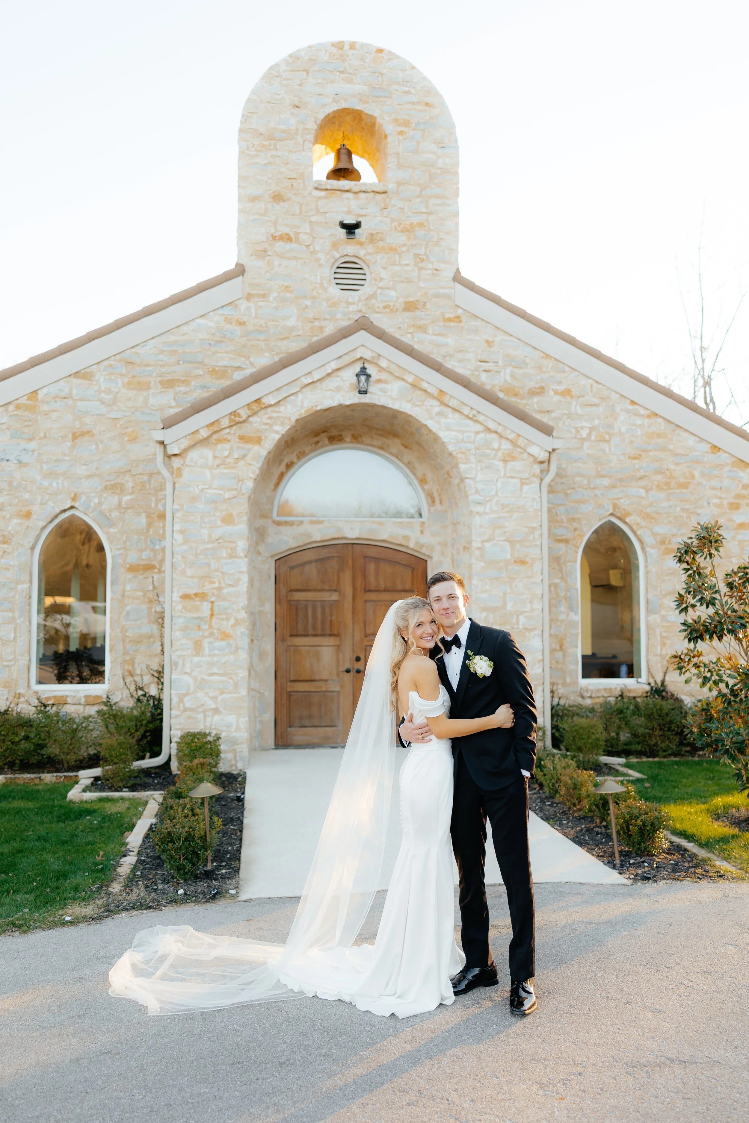 A bride and groom stand together outside a stone church during their wedding, smiling and embracing, with the church's wooden doors and stone architecture in the background.