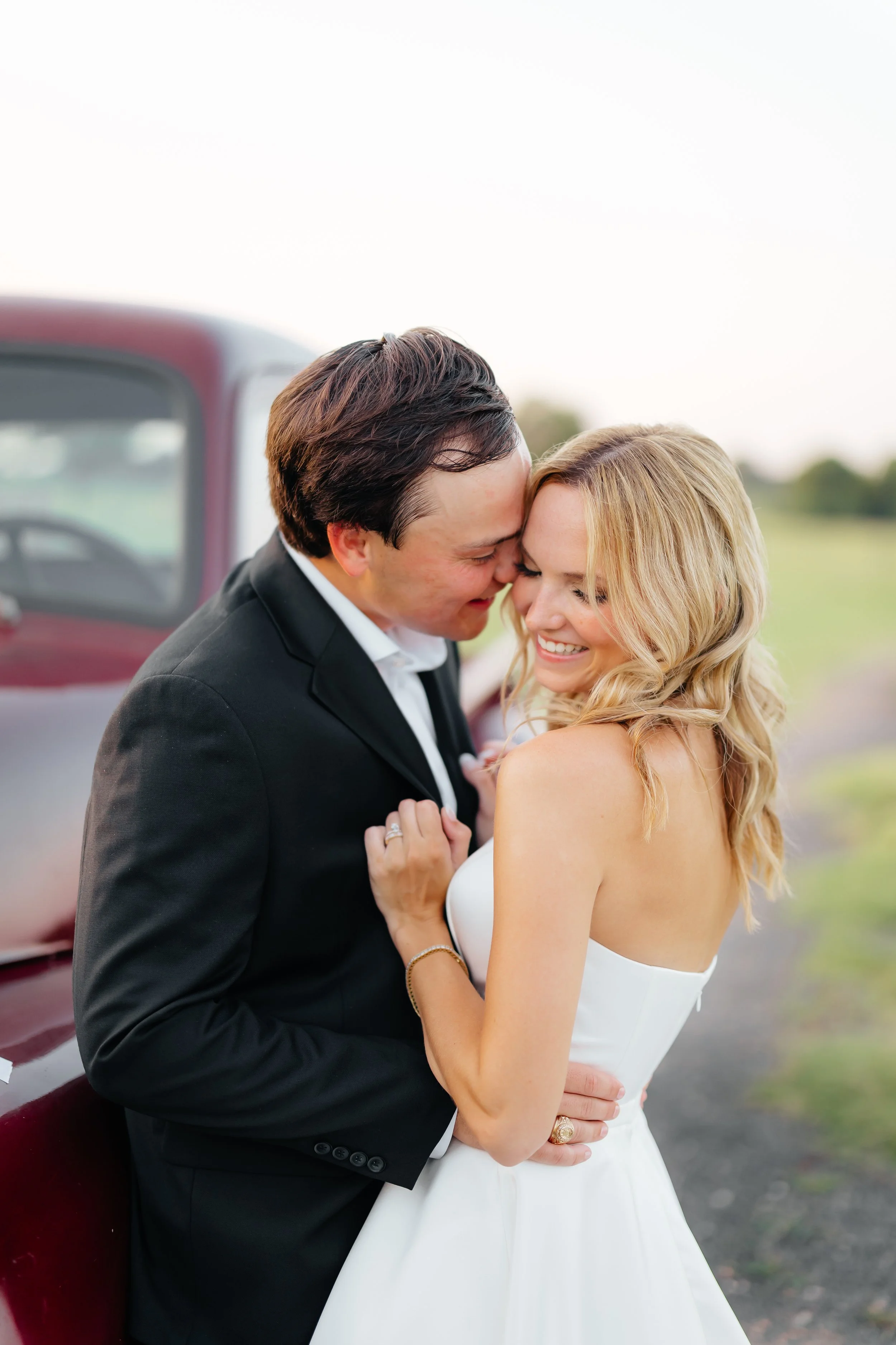A newlywed couple about to kiss outdoors near a vintage car, with the groom in a black suit and the bride in a strapless white wedding dress, smiling and appearing happy.