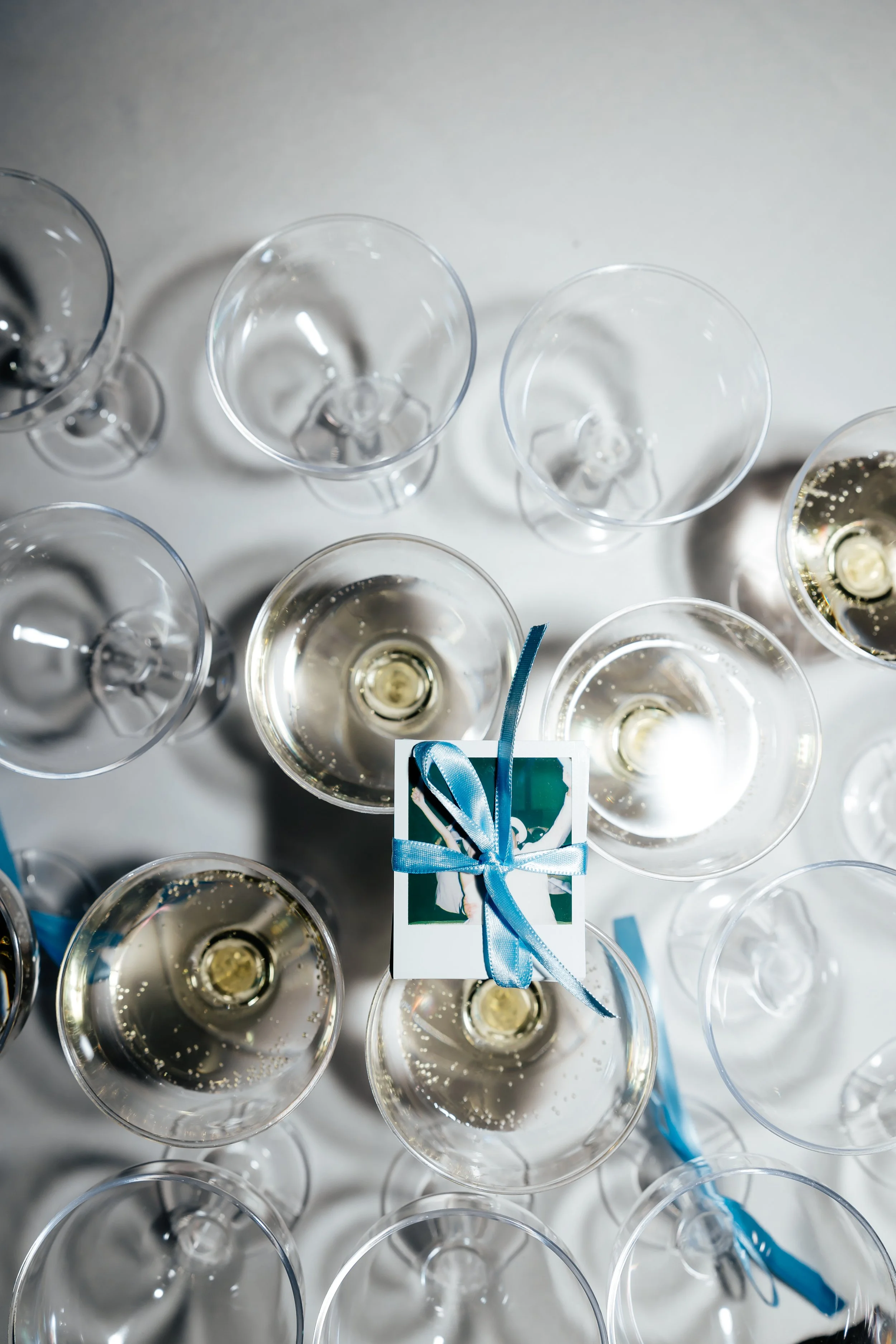 Multiple empty and filled wine glasses with a small gift box tied with a blue ribbon on a white surface, viewed from above.
