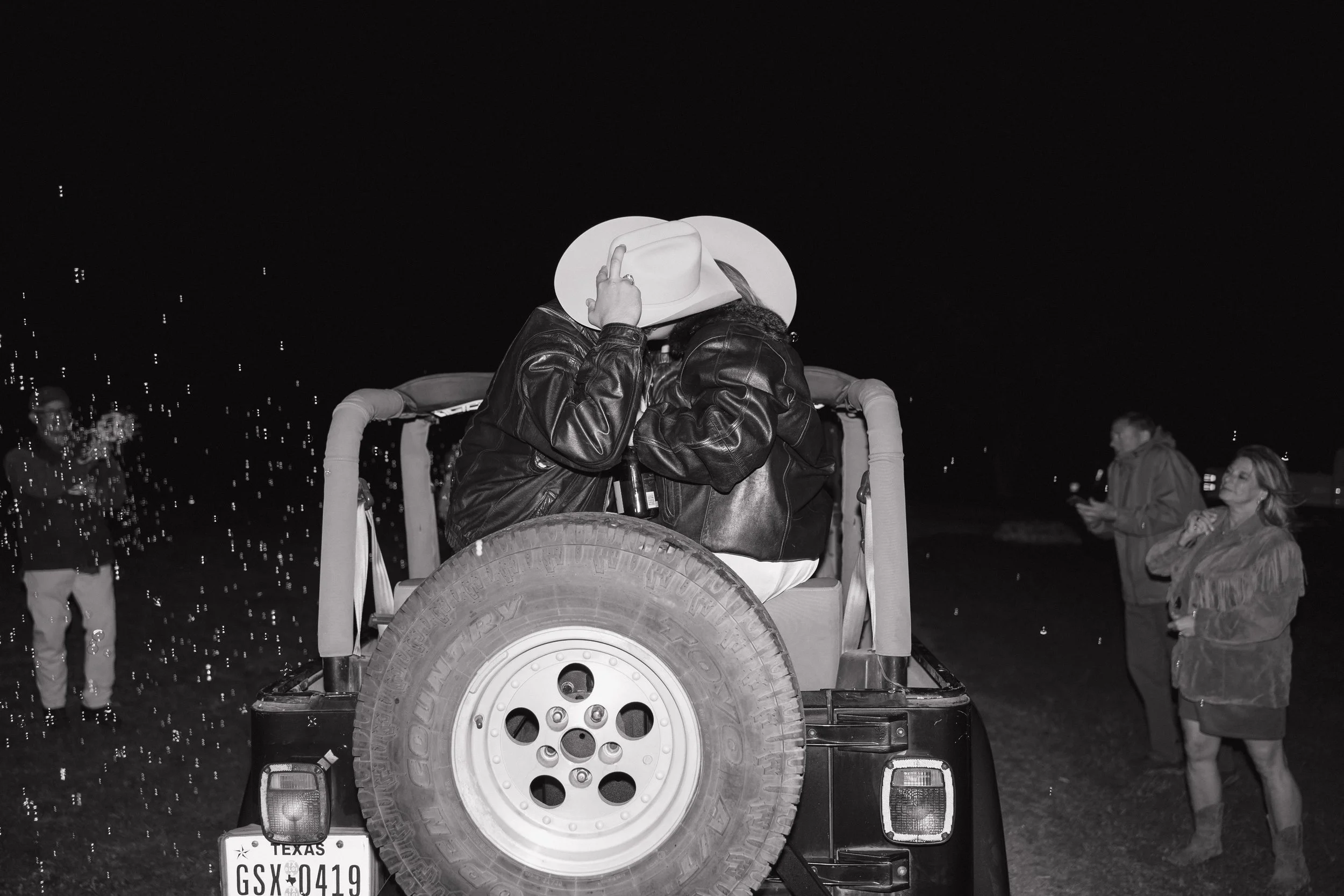 Two people wearing leather jackets and a cowboy hat sitting on the back of a Jeep, with friends standing nearby at night.