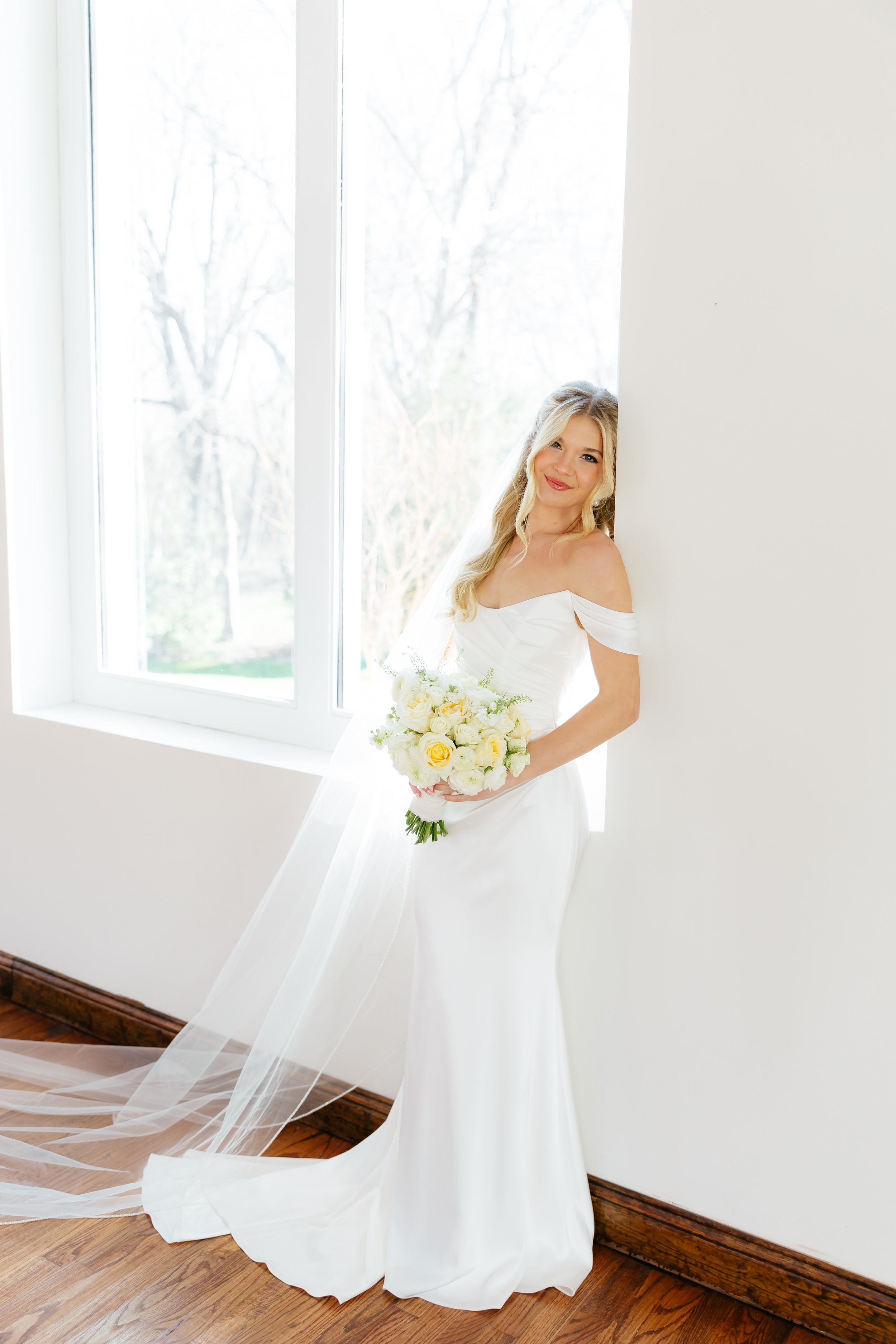 A bride in a white wedding gown holding a bouquet of white and yellow roses, standing by a large window with trees outside.