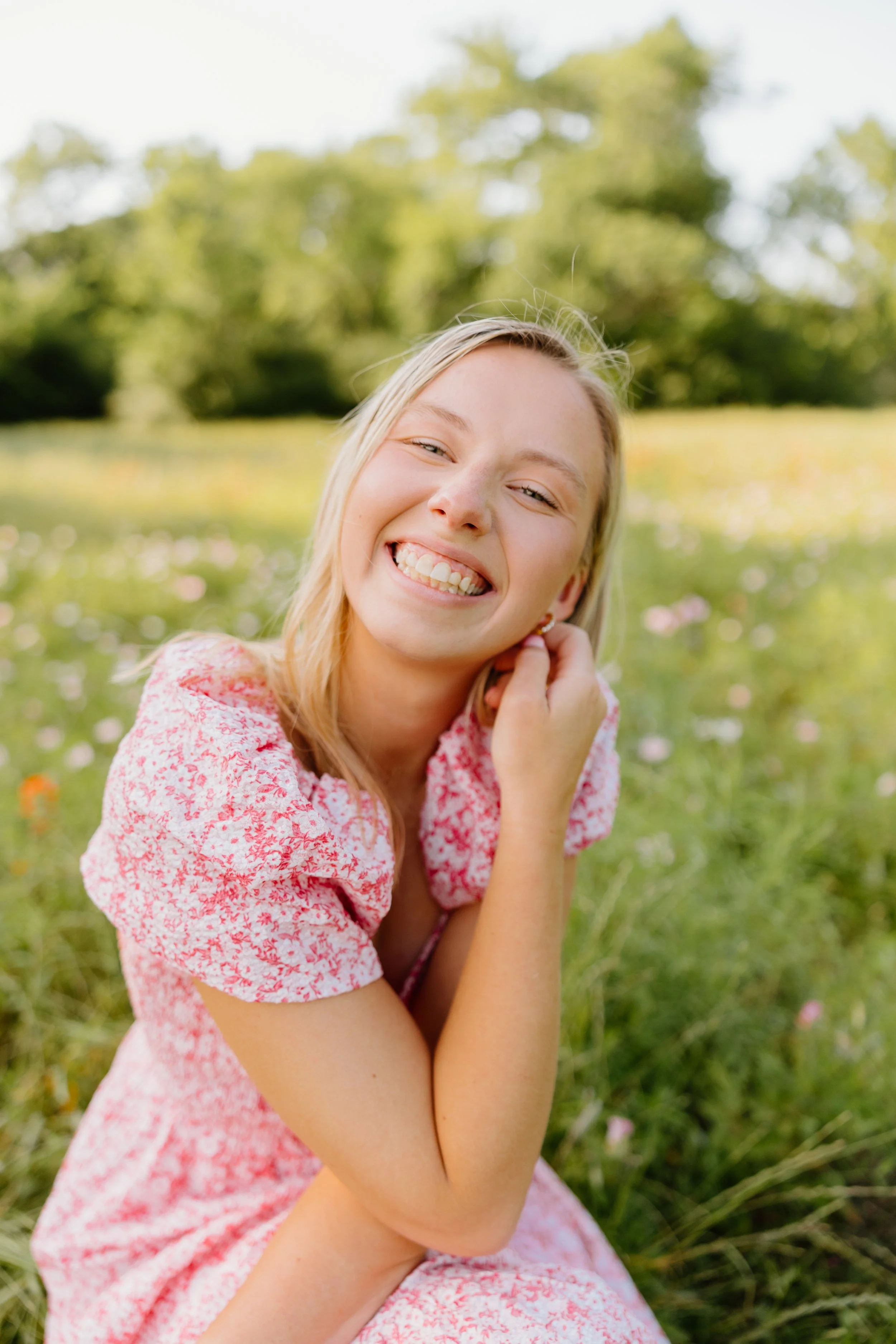A young woman smiling and sitting in a field of flowers and grass with trees in the background.