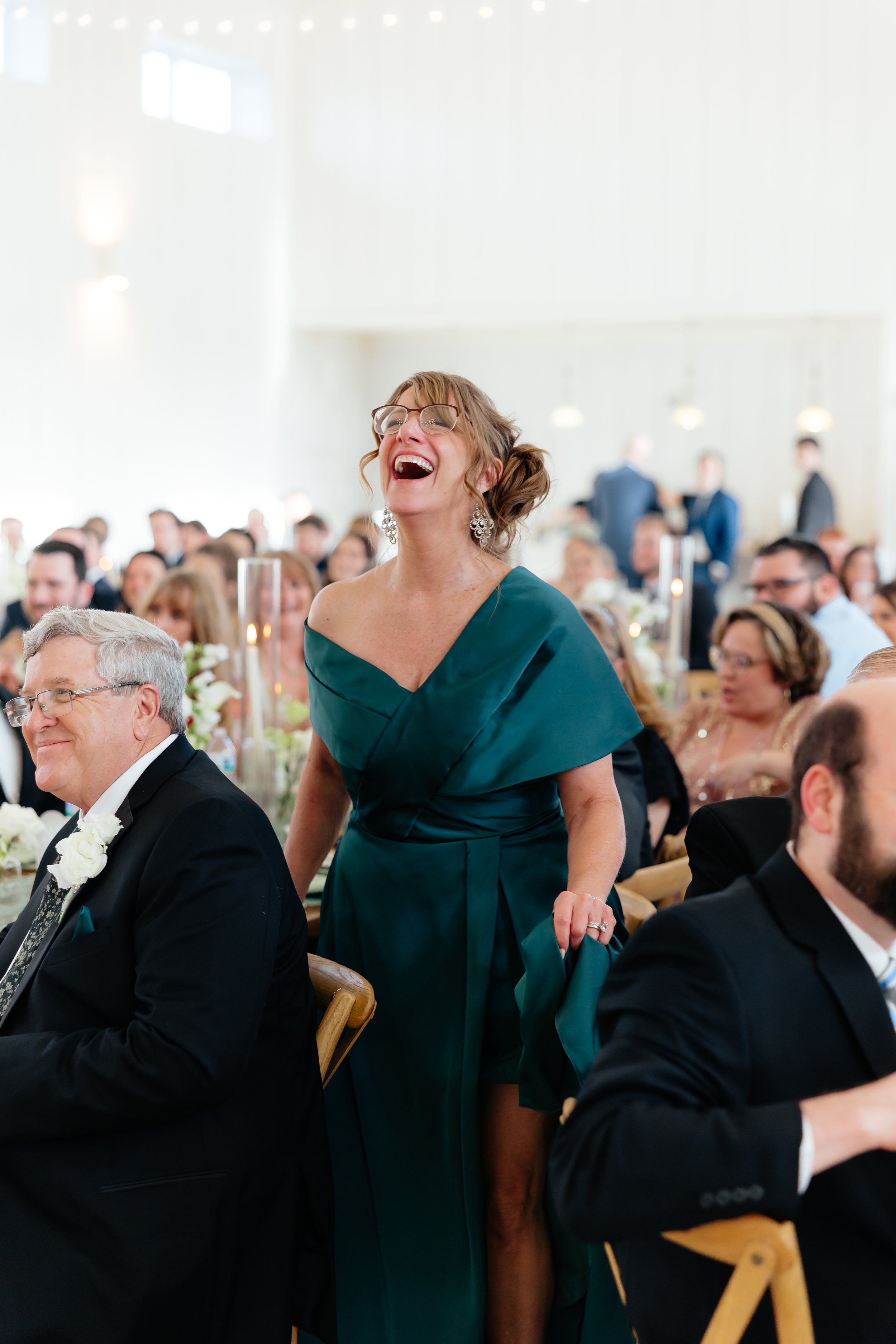 A woman in a teal dress and glasses laughing at a wedding reception, surrounded by seated guests in formal attire.