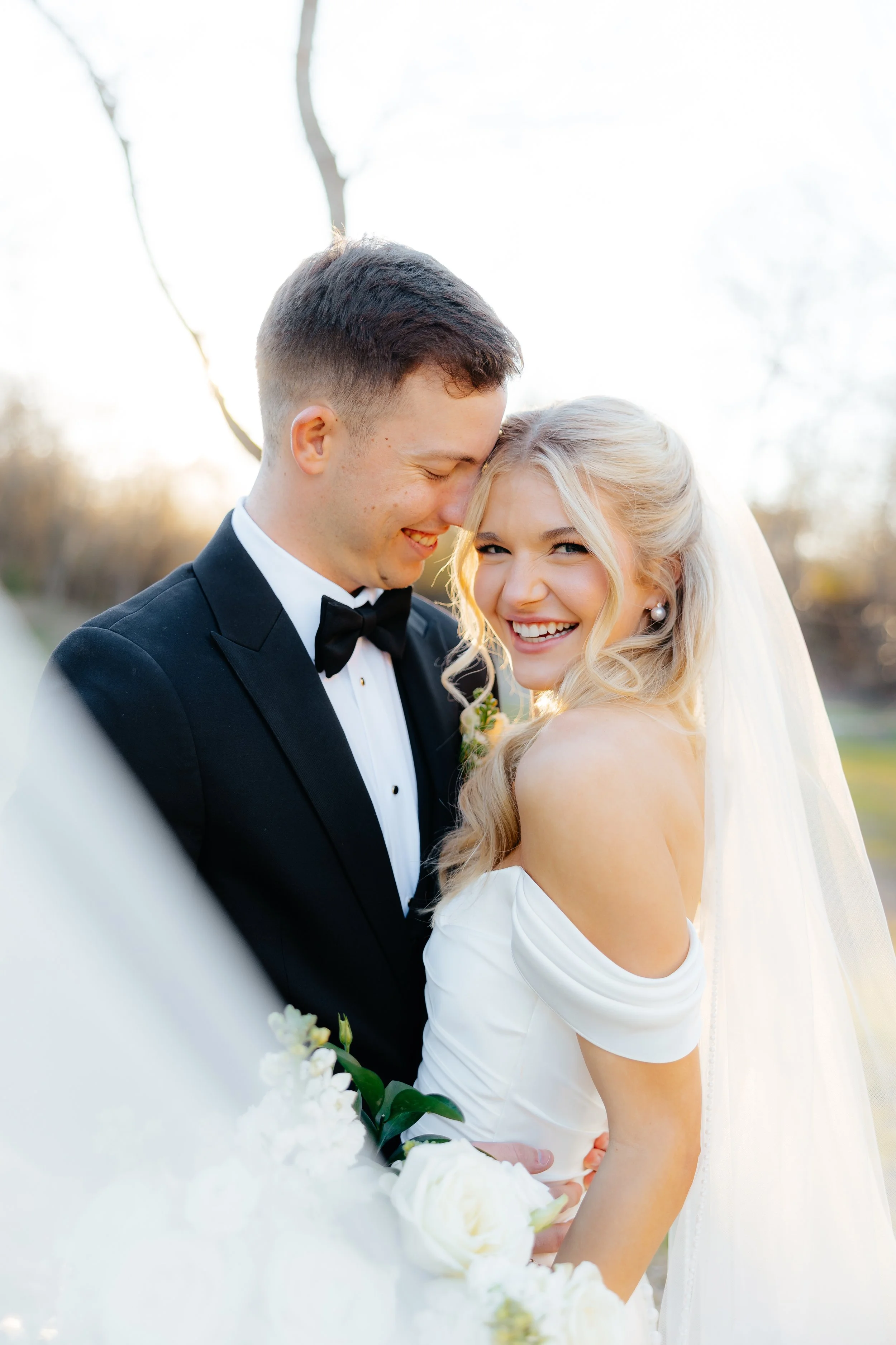 A newlywed couple smiling and laughing outdoors during daytime, with the groom in a black tuxedo and the bride in a white wedding dress, holding a bouquet of flowers.