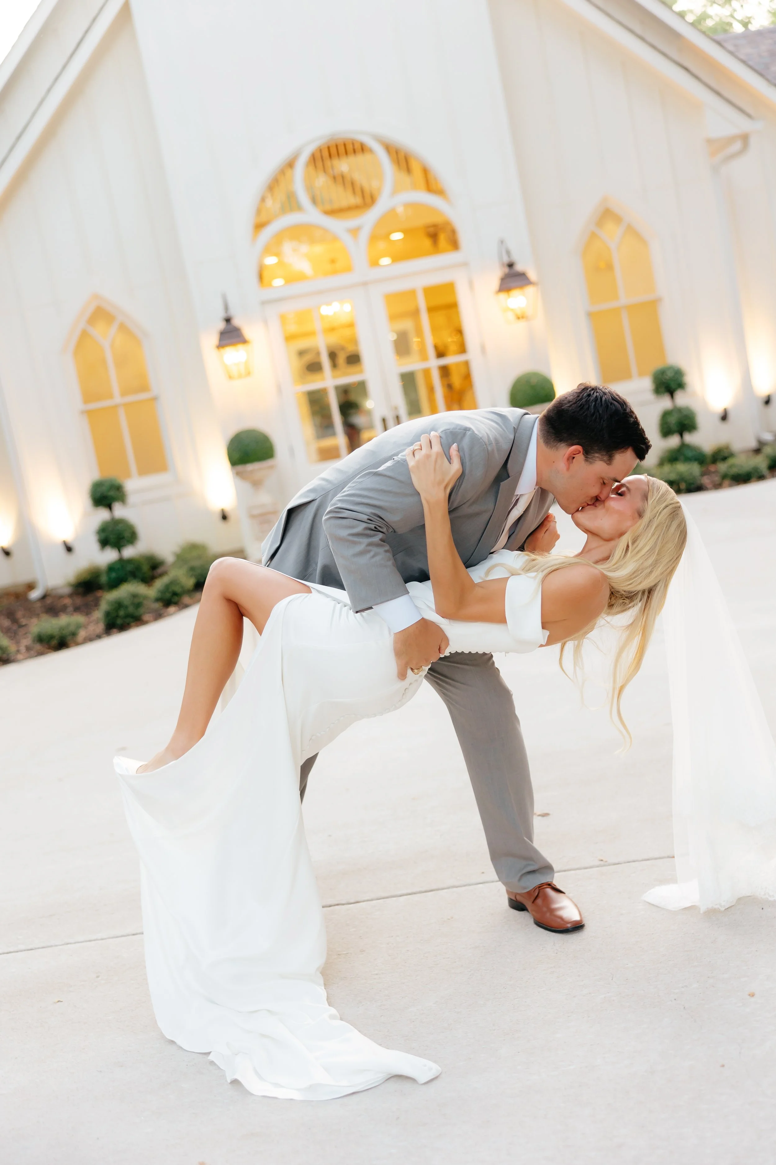 A newlywed couple sharing a kiss outside a white church at sunset. The groom in a gray suit dips the bride in a white wedding gown.