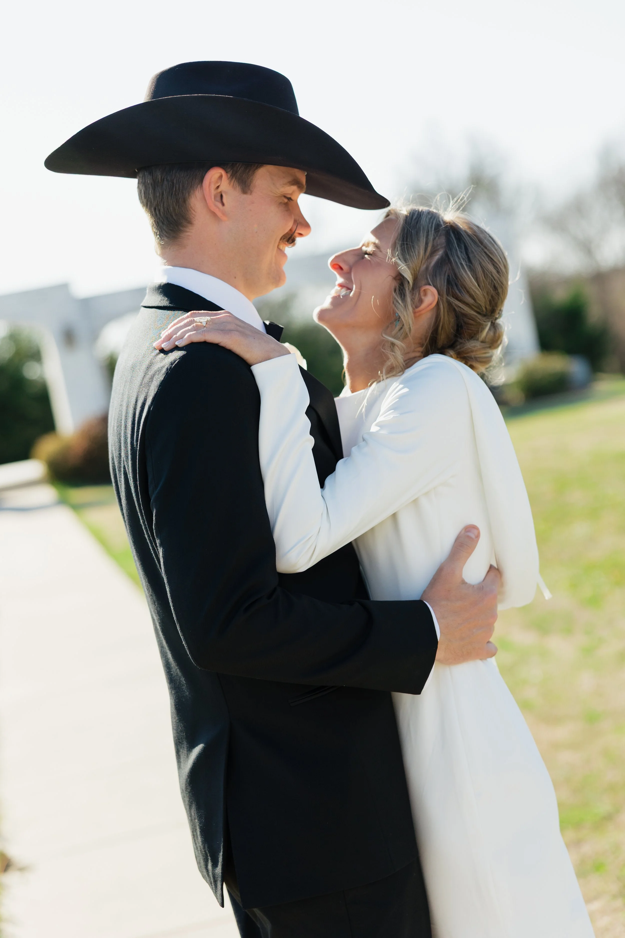 A couple dressed in wedding attire sharing a joyful moment outdoors, with the man wearing a black suit and large black hat and the woman in a white dress.