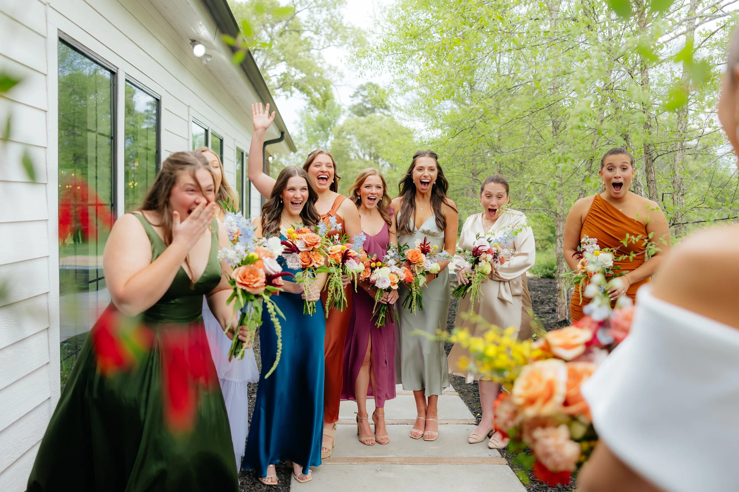 A group of women in colorful dresses holding bouquets, appearing joyful and excited as they greet someone, likely at a wedding or celebration outdoors.