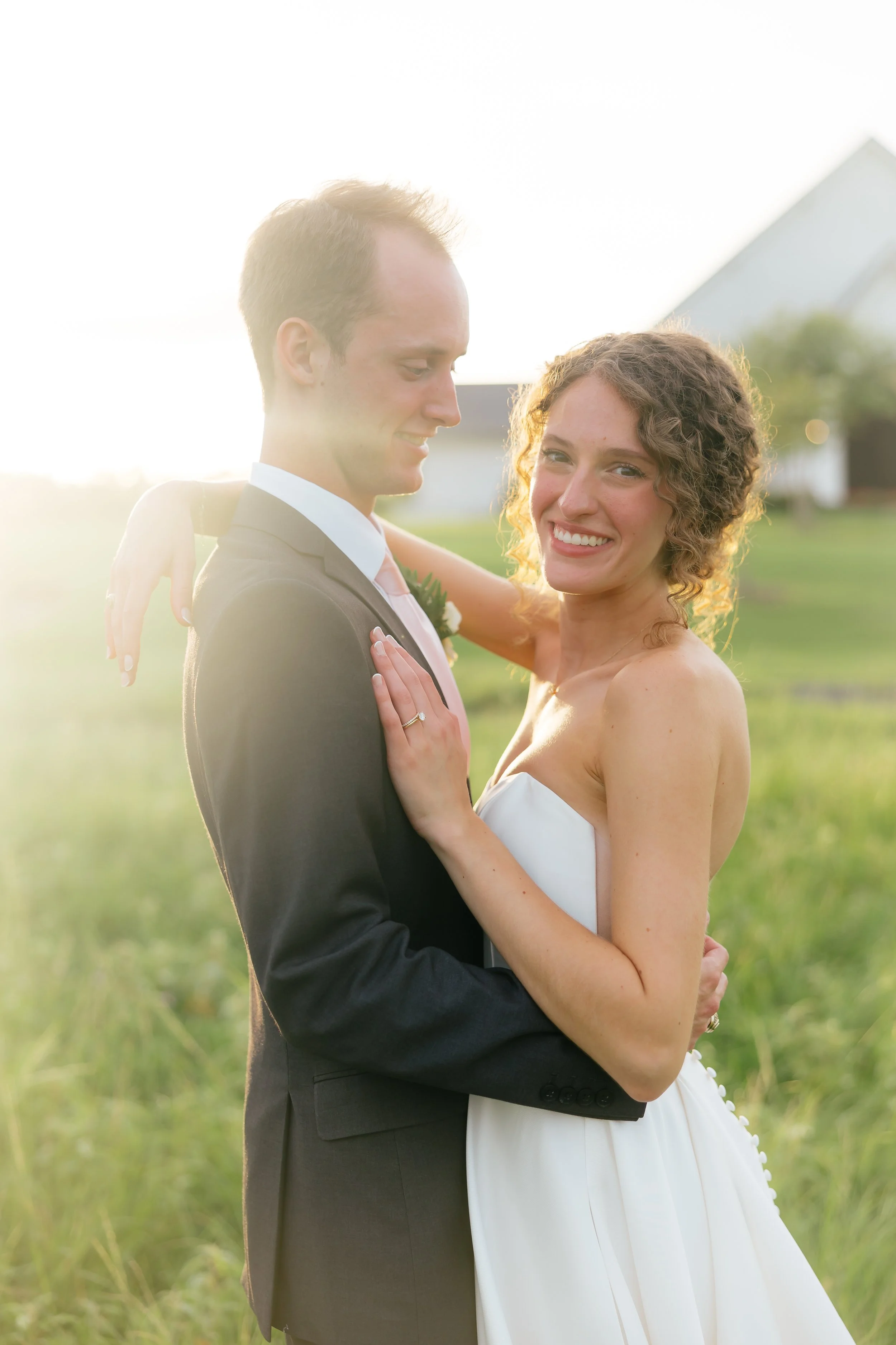 A newlywed couple embracing outdoors during sunset, with a white barn in the background.