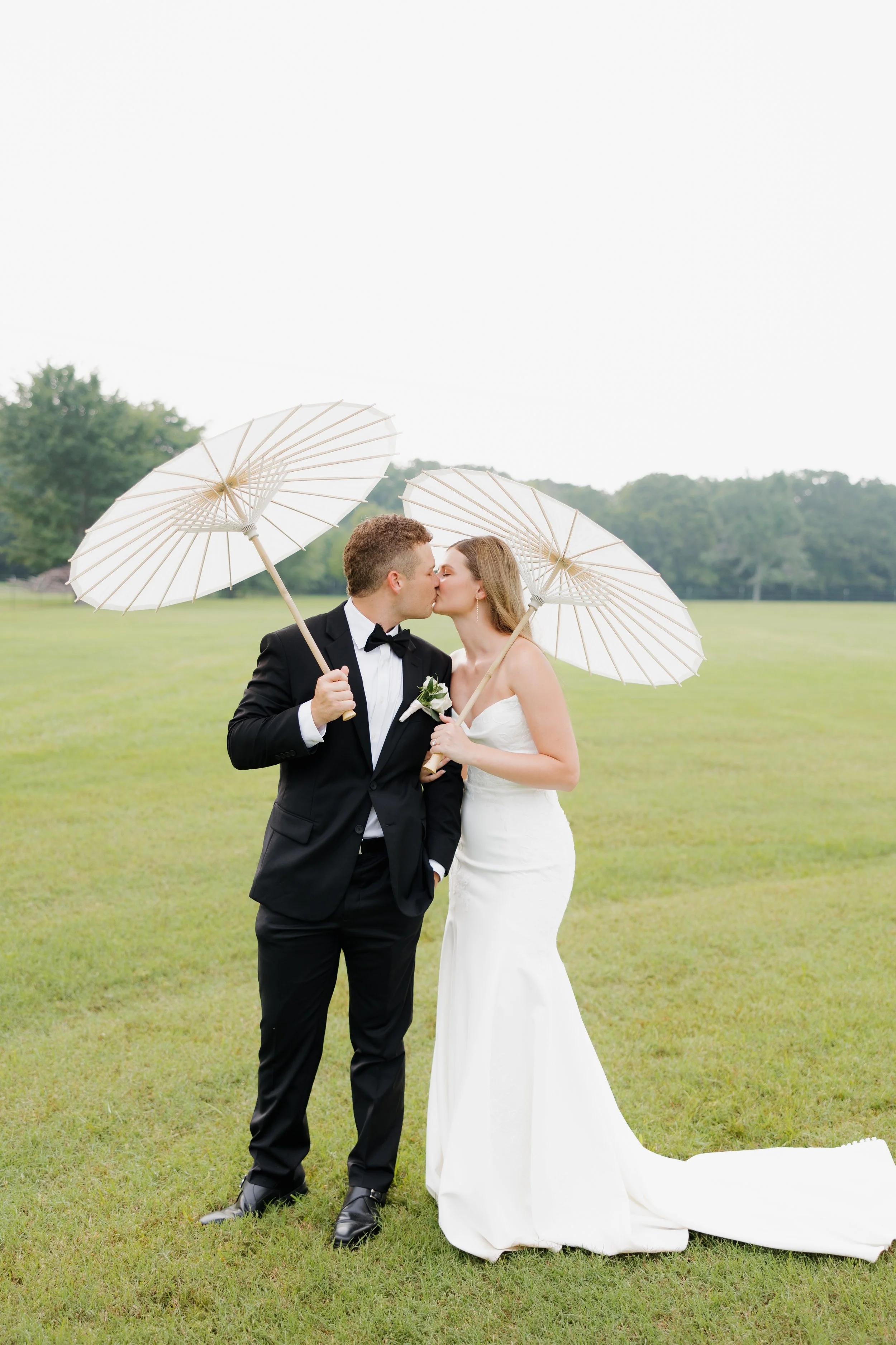 A bride and groom, dressed in wedding attire, are kissing outdoors on a grassy field, holding white parasols.
