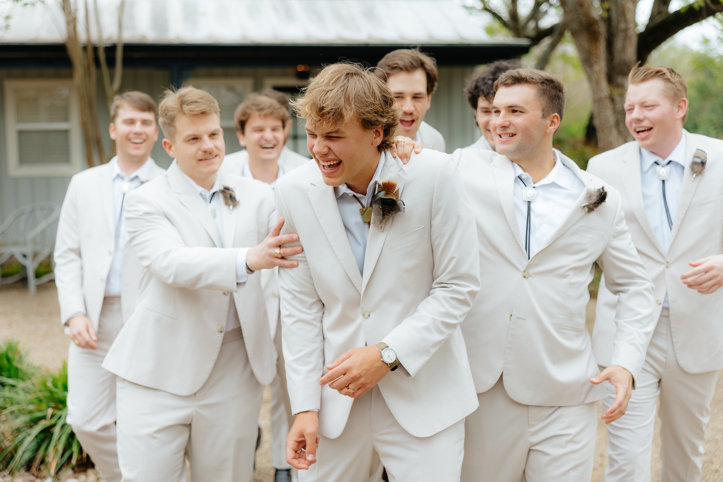 Group of men dressed in white suits, laughing and smiling outdoors, with one man in the center showing a beard and a watch, surrounded by others with bows and boutonnières.