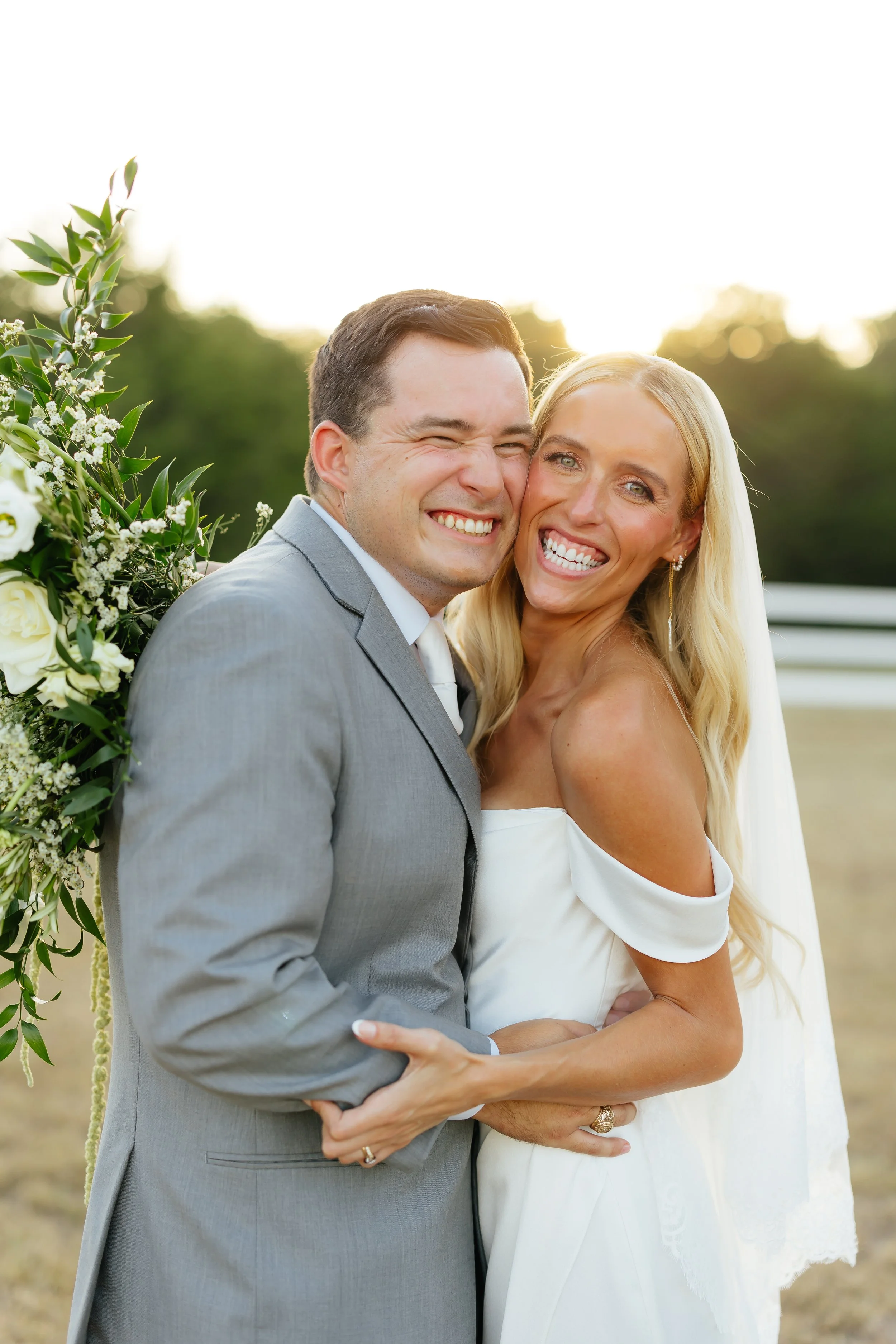 A newlywed couple smiling and hugging outdoors during sunset, with a floral arch in the background.