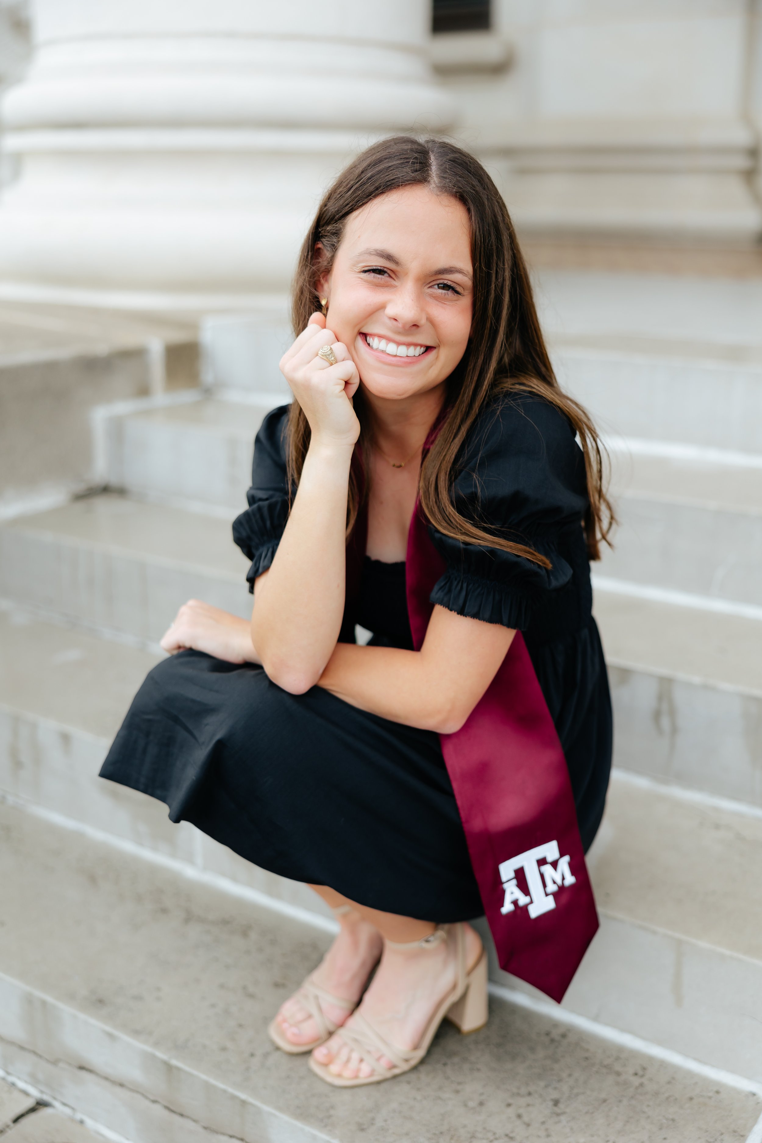 Young woman in black dress with maroon sash sitting on steps, smiling at camera.