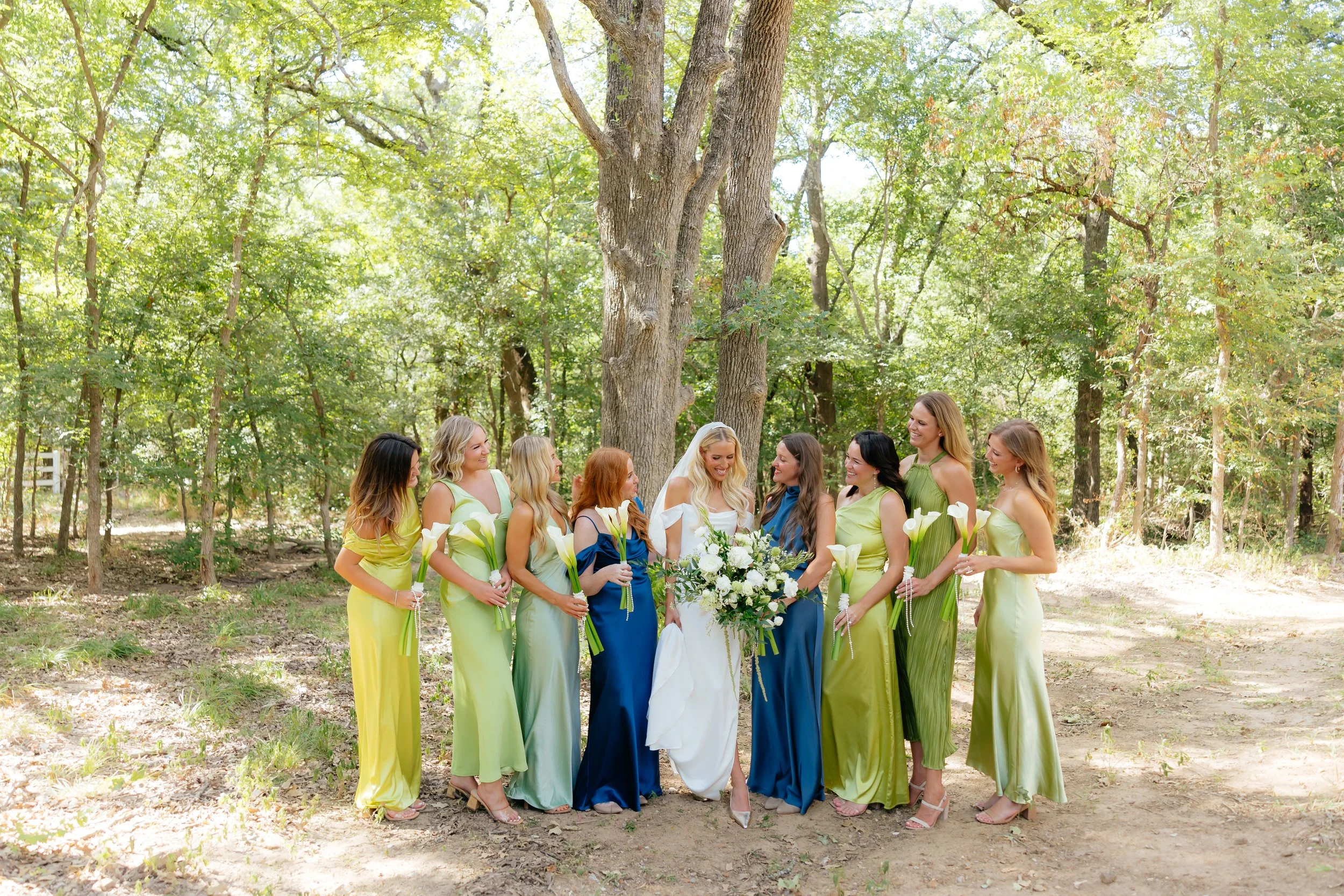 A bride in white wedding dress with a large bouquet stands surrounded by ten women in colorful dresses, all holding calla lilies, outdoors in a forested area with tall trees and green foliage.
