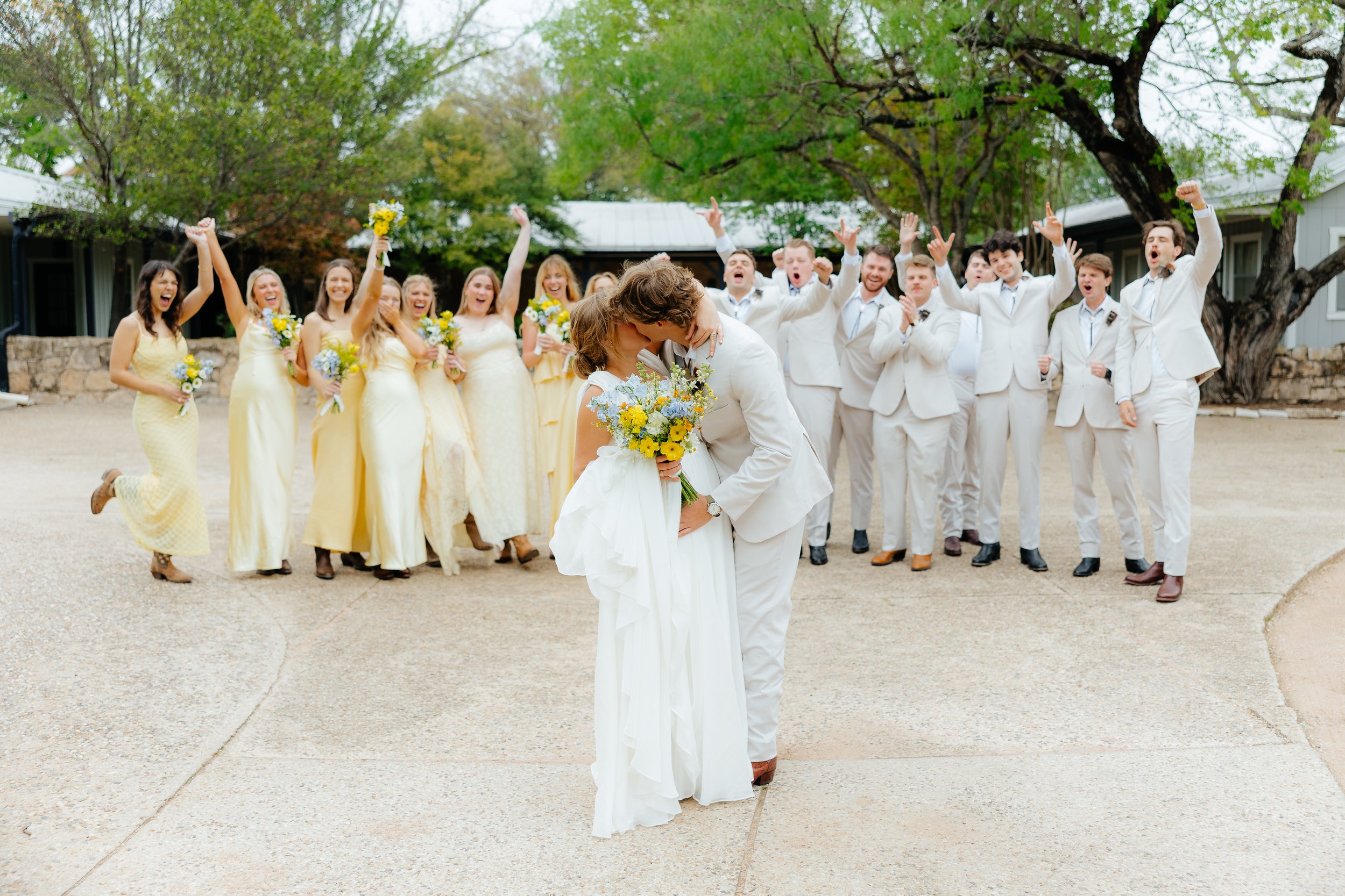 A wedding party outdoors with the bride and groom in the foreground sharing a kiss, surrounded by bridesmaids and groomsmen celebrating and cheering.