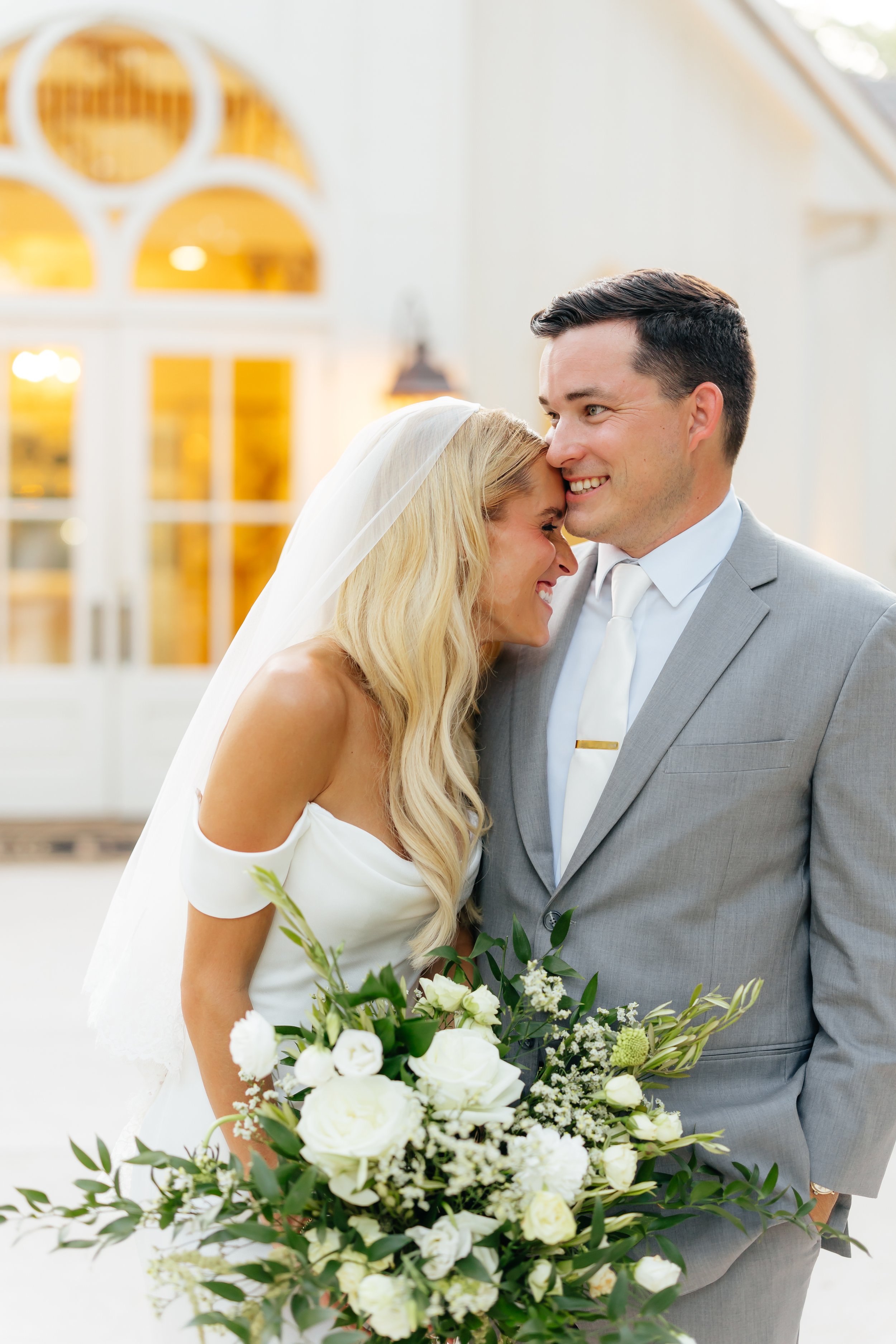 A bride and groom share a joyful moment on their wedding day, with the bride holding a bouquet of white flowers and greenery, indoors with bright natural light.