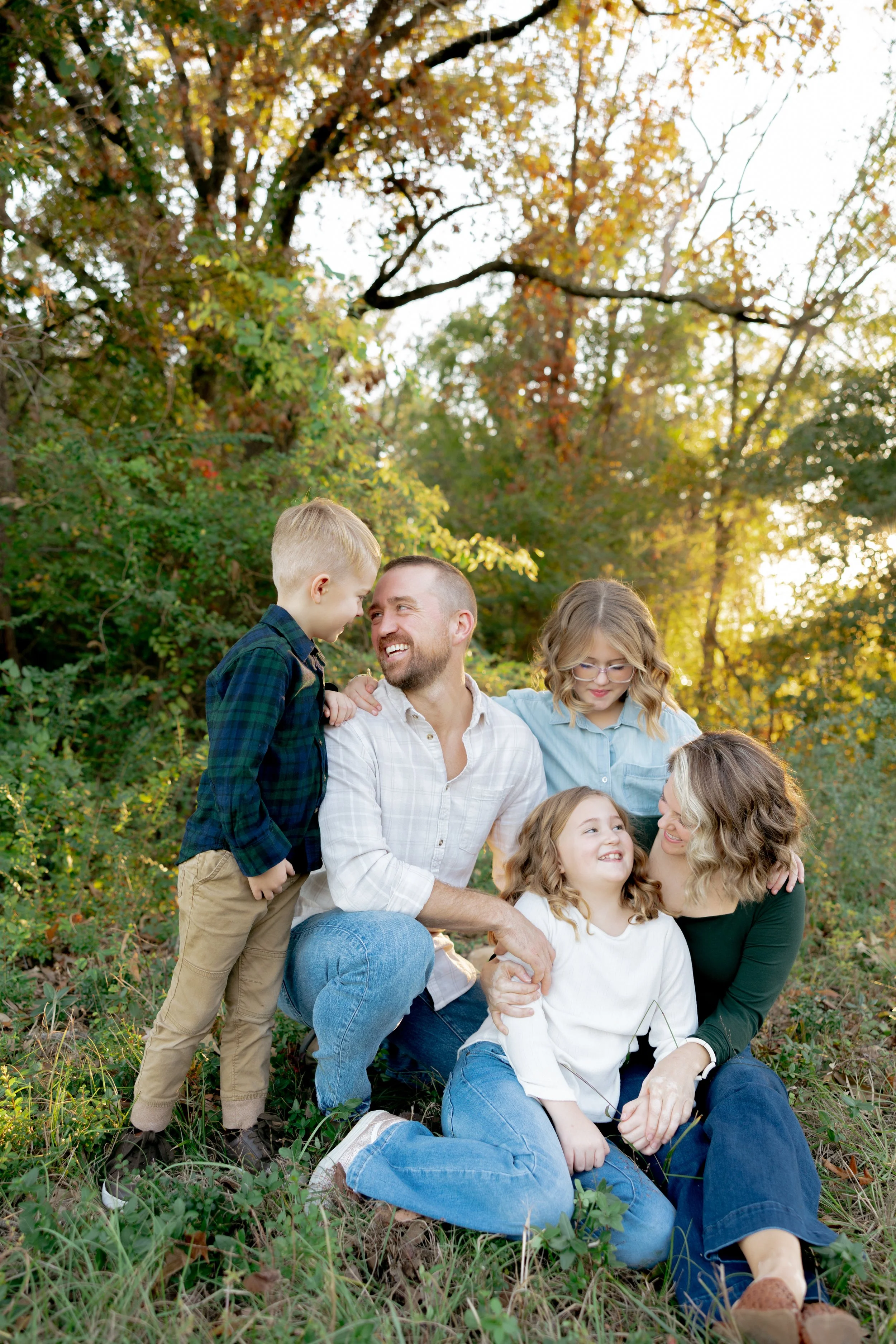 A family of six enjoying time outdoors in a park during autumn, sitting on the grass with trees and sunlight in the background.