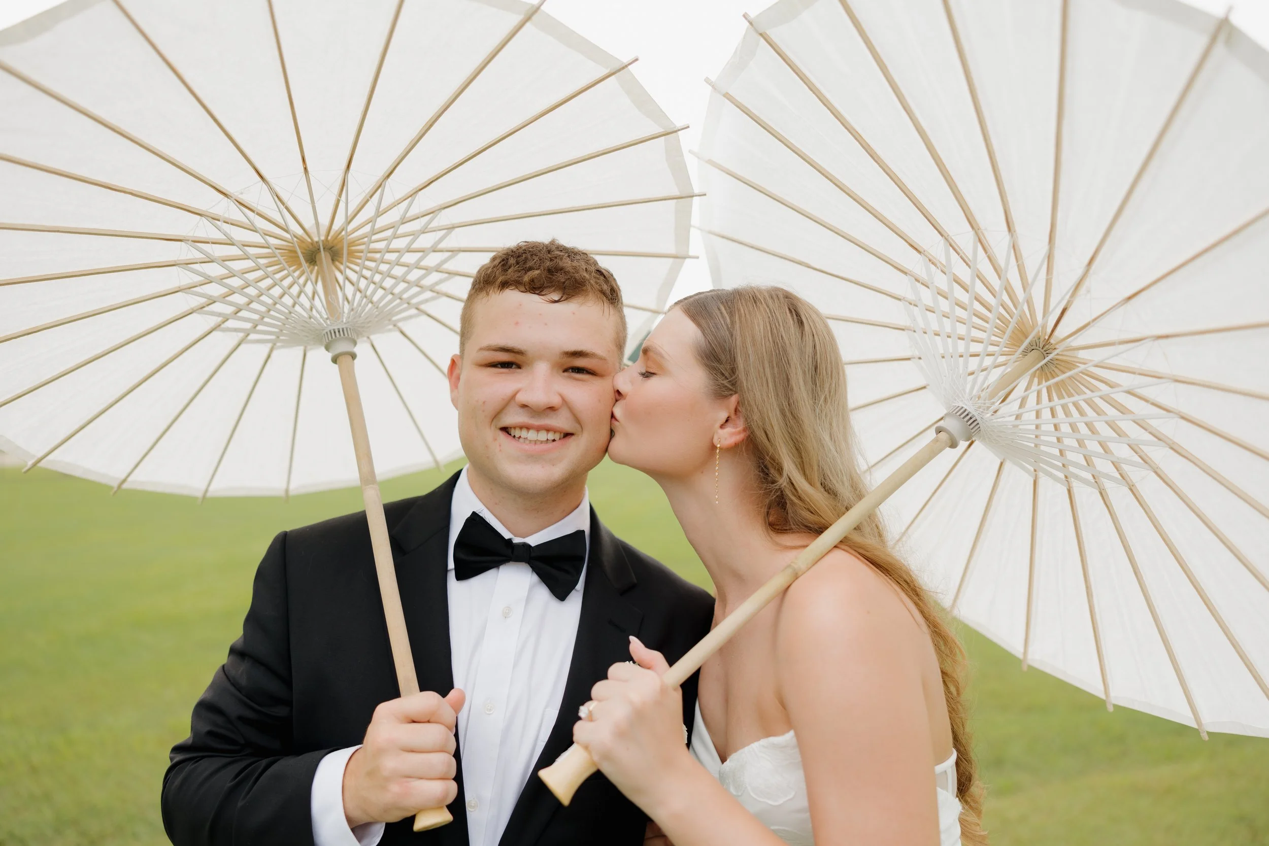 A bride and groom in wedding attire sharing a kiss under white umbrellas outdoors.