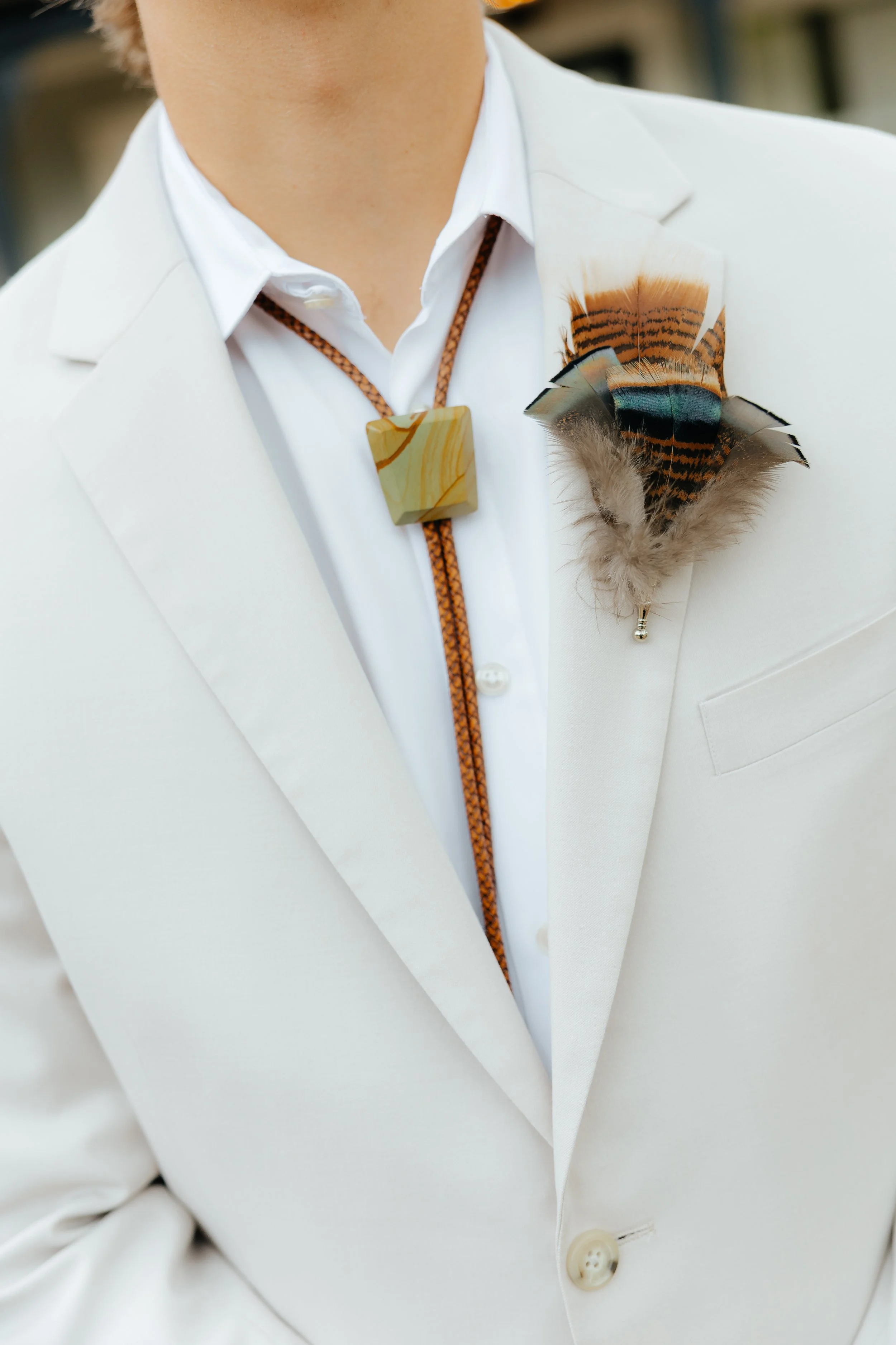 Close-up of a person wearing a white suit, white shirt, with a feathered and beaded accessory on their lapel, and a necklace with a square pendant.