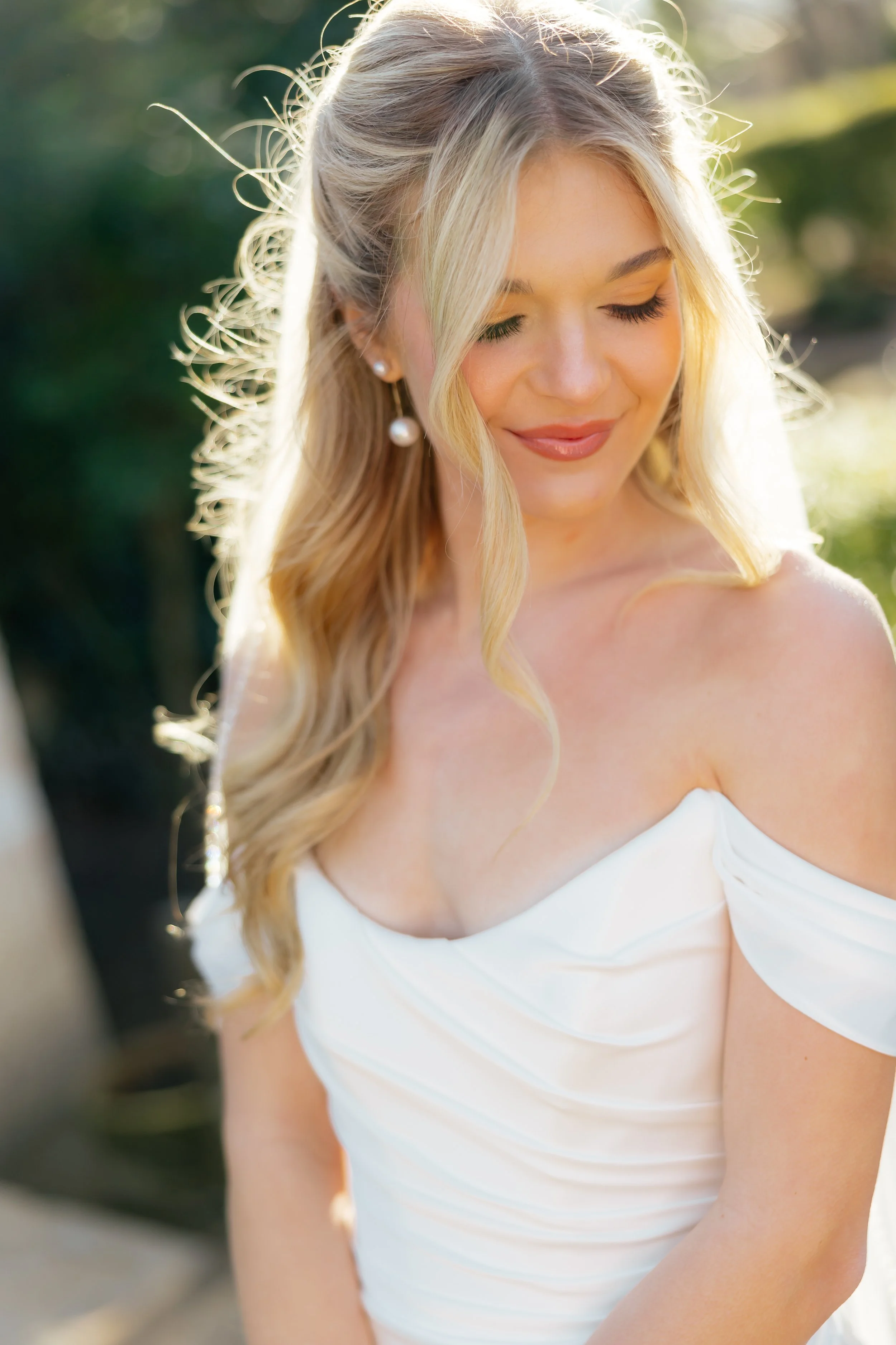 A woman wearing a white off-shoulder dress with long blonde hair, pearl earrings, and makeup, standing outdoors with sunlight creating a soft glow.