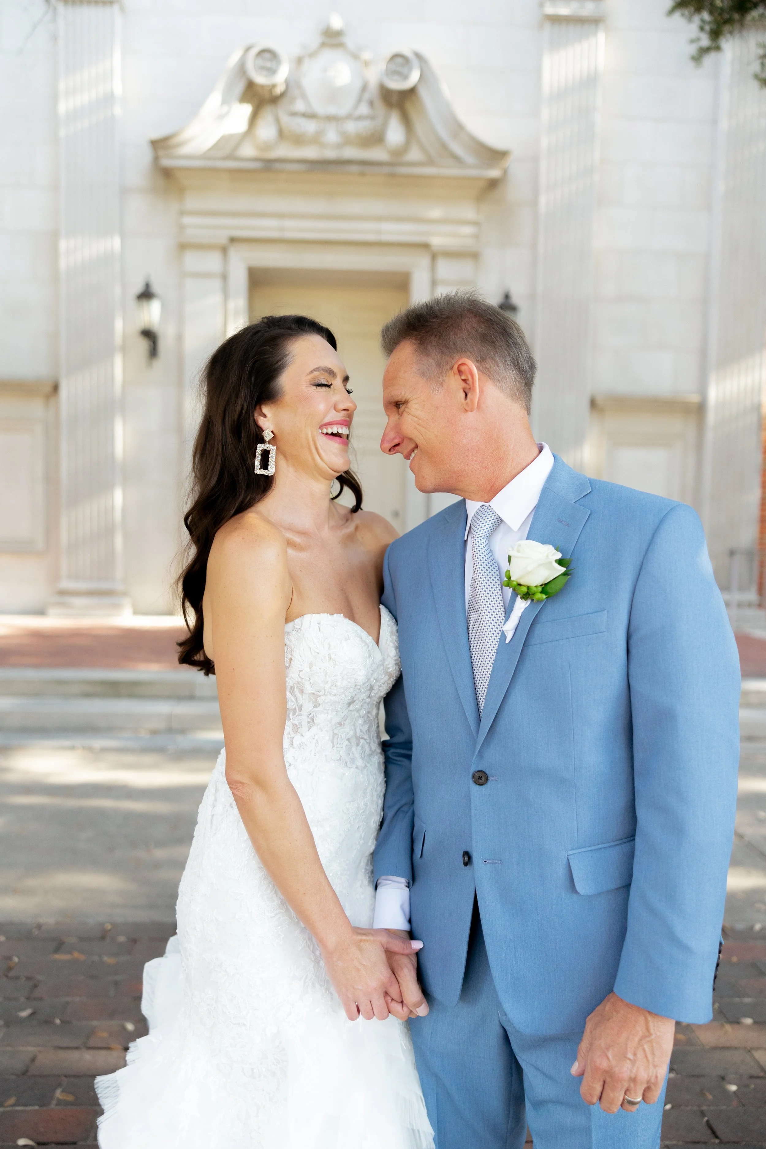 A bride and groom holding hands and smiling at each other in front of a building with columns, during their wedding.