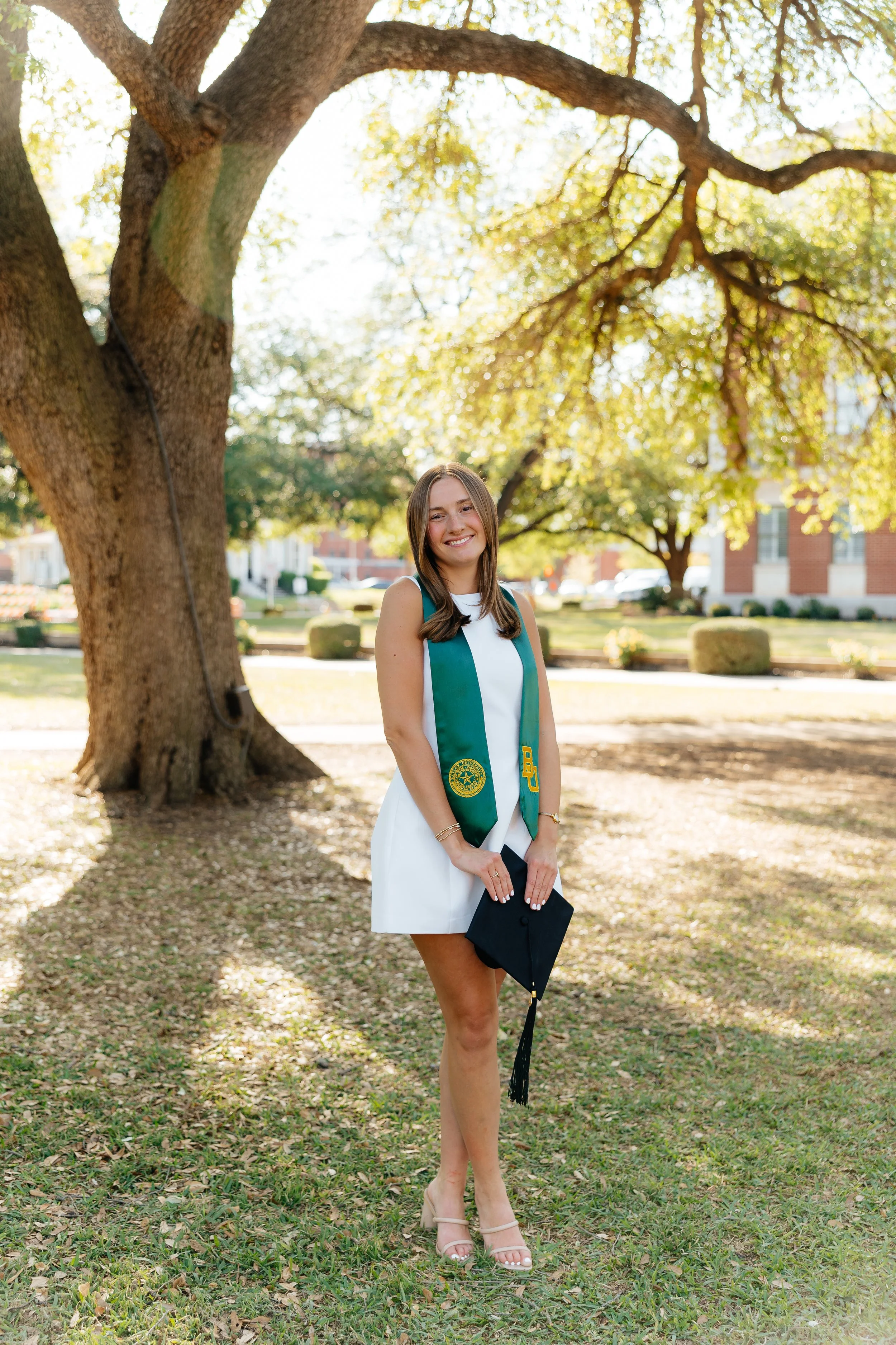A young woman in a graduation cap and gown holding her cap in her hand, standing outdoors near a large tree with yellow leaves, smiling.