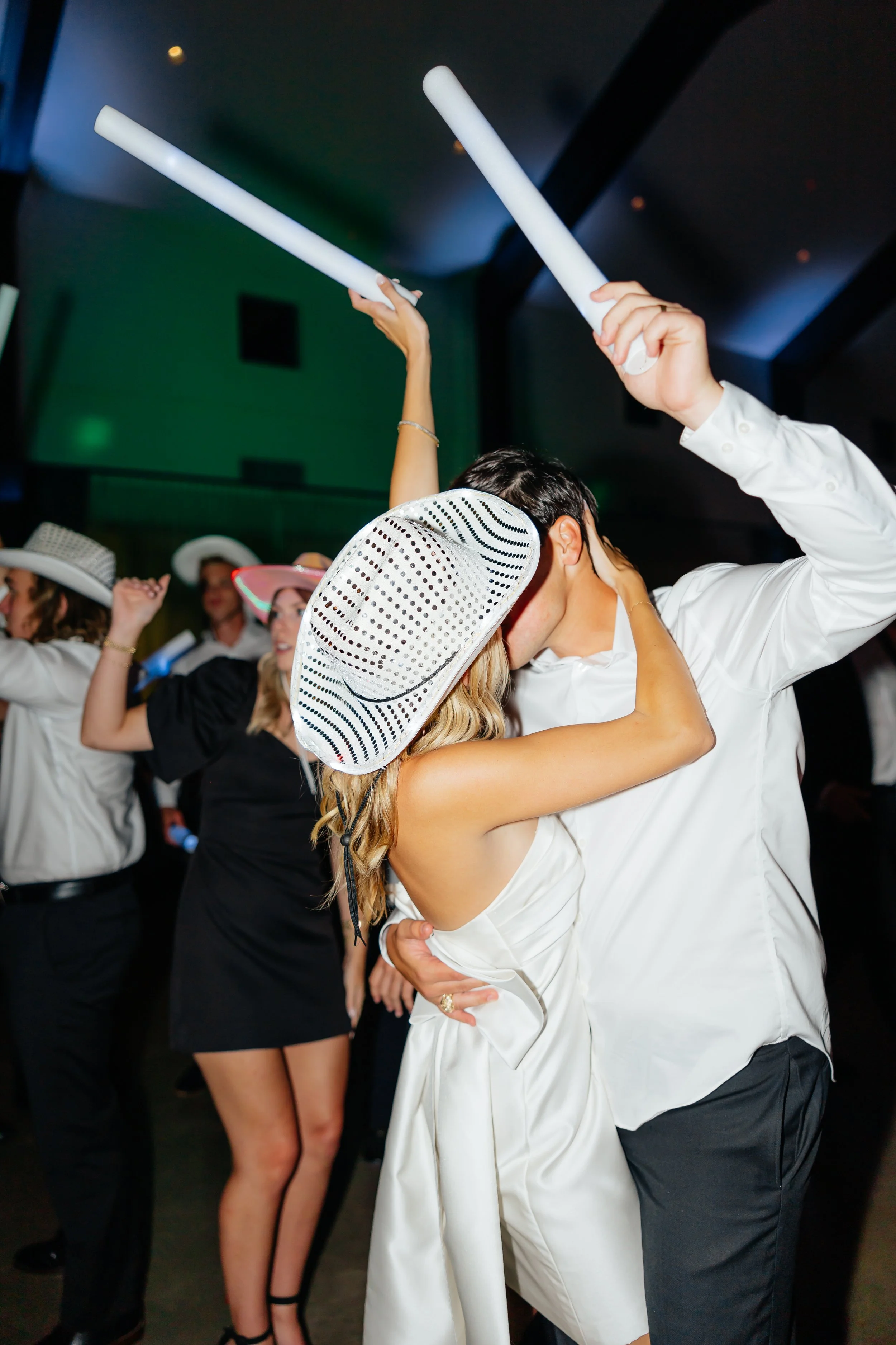 Couple dancing closely at a celebration wearing white and black attire, with women in the background wearing hats and smiling.