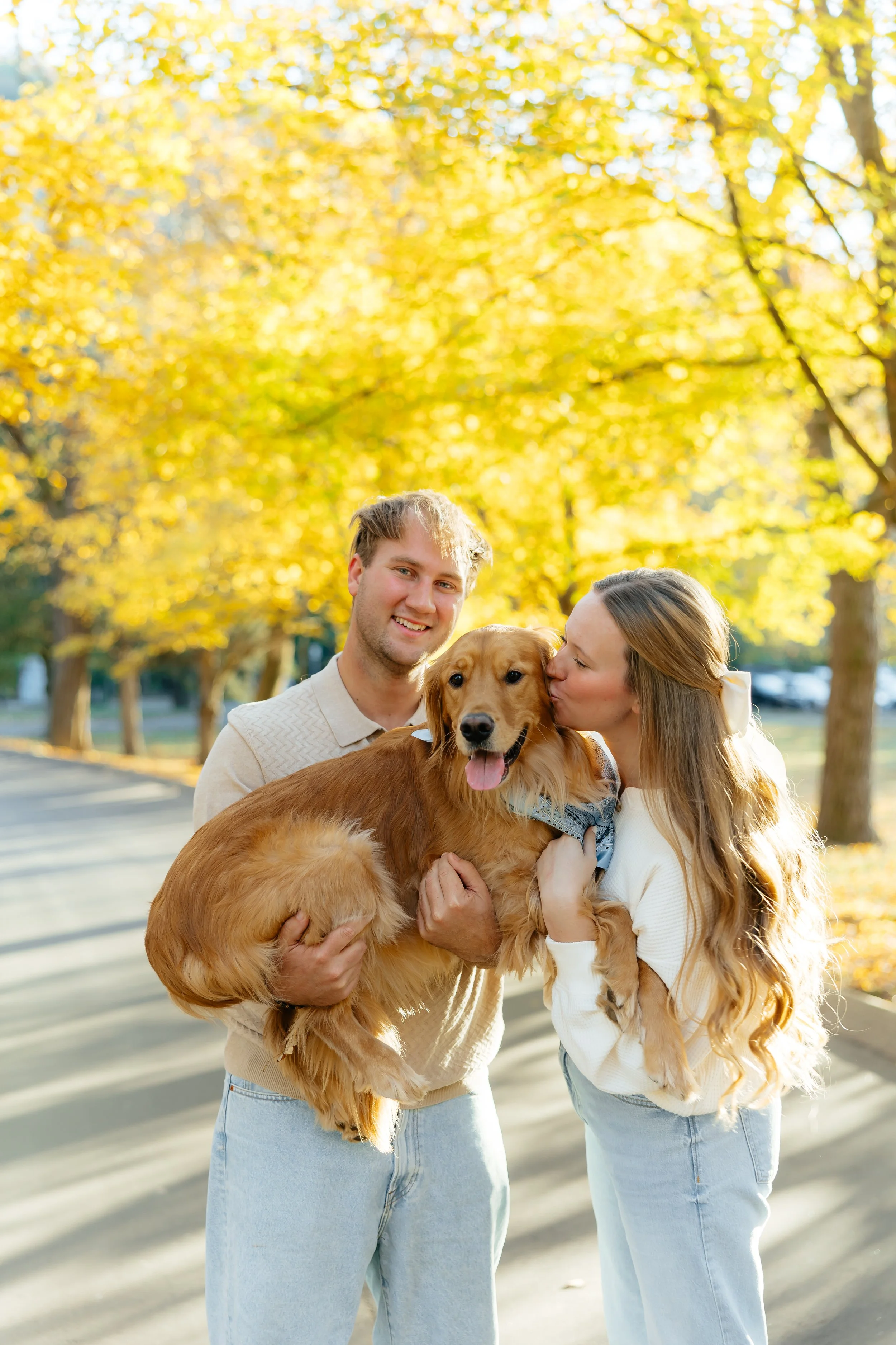 A young couple with a golden retriever dog outdoors during autumn, with golden leaves on trees in the background. The woman is kissing the dog on the cheek, and the man is smiling at the camera while holding the dog.