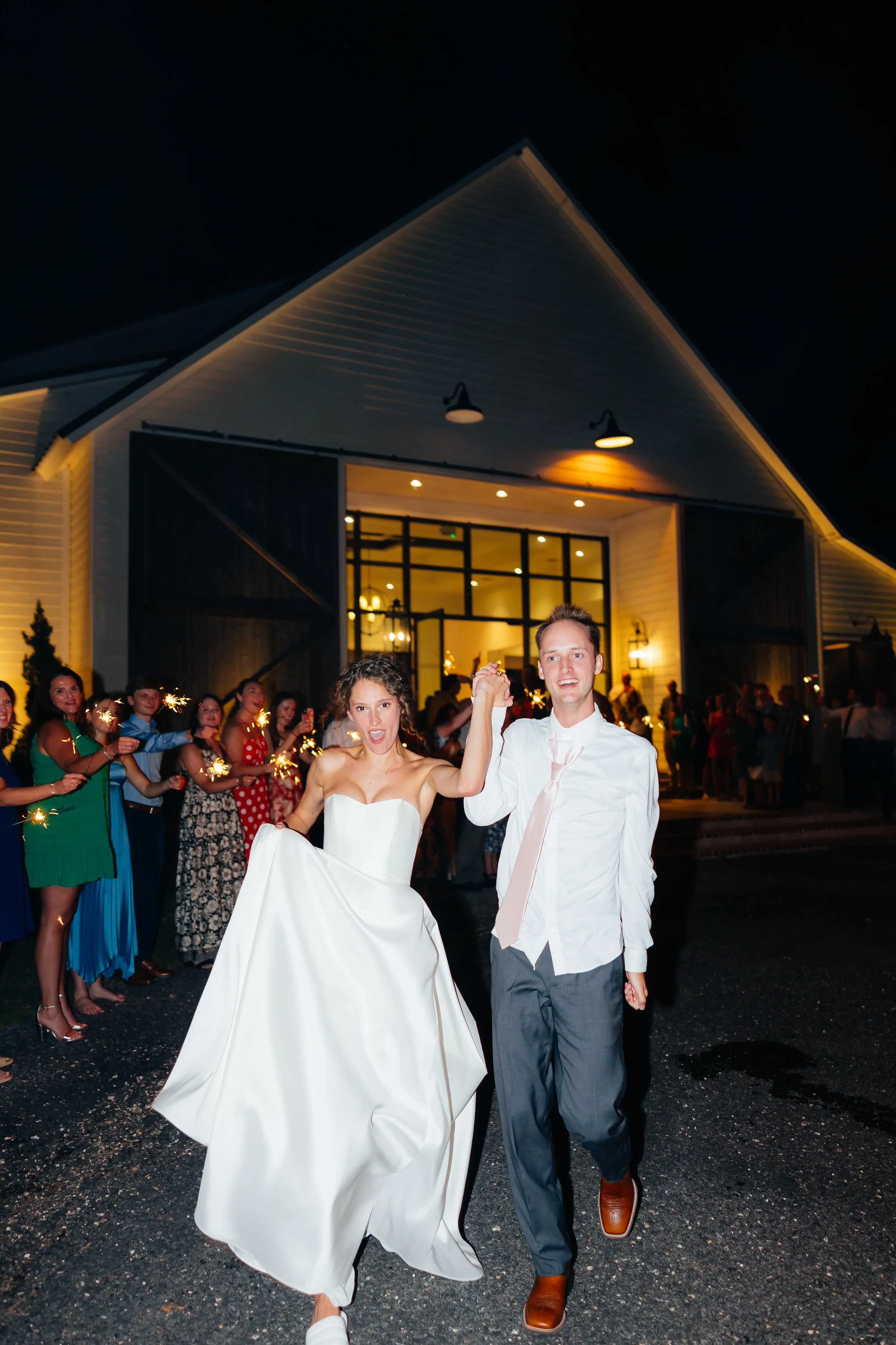 A newlywed couple holding hands and celebrating at their wedding reception outside a lit barn at night while guests hold sparklers in the background.