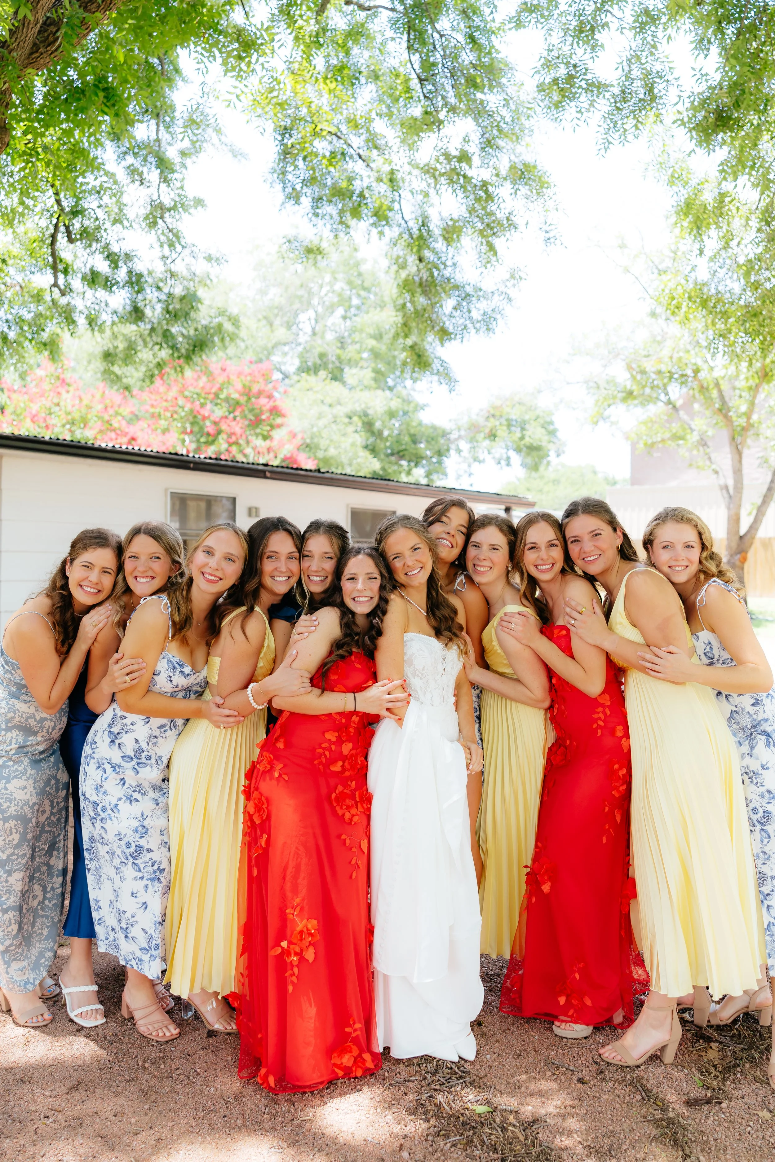 Group of women dressed in colorful dresses, smiling and hugging outdoors under trees.