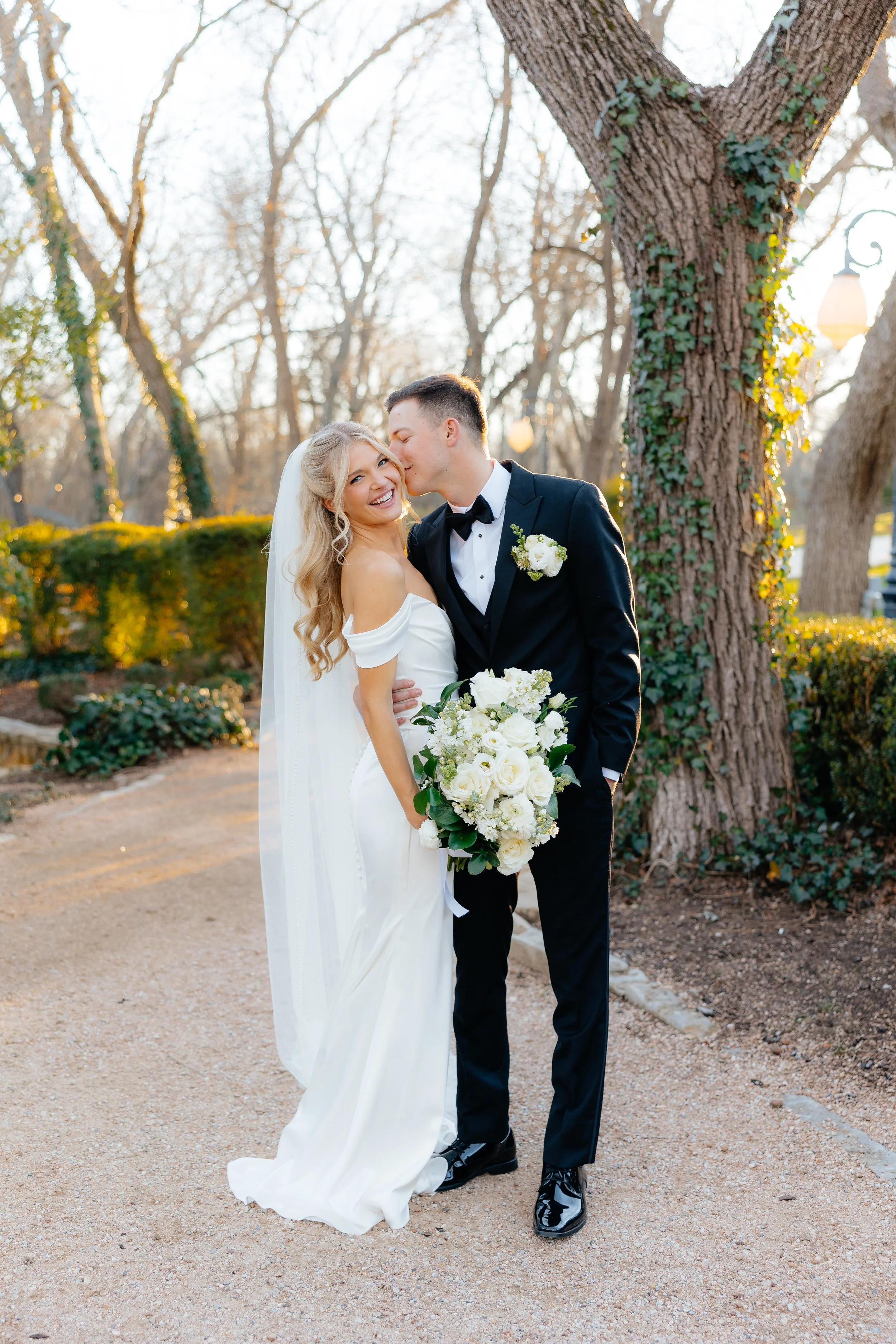 Bride and groom on their wedding day, standing outdoors with trees and shrubs in the background, the bride holding a bouquet of white flowers, the groom kissing her on the cheek, during sunset.