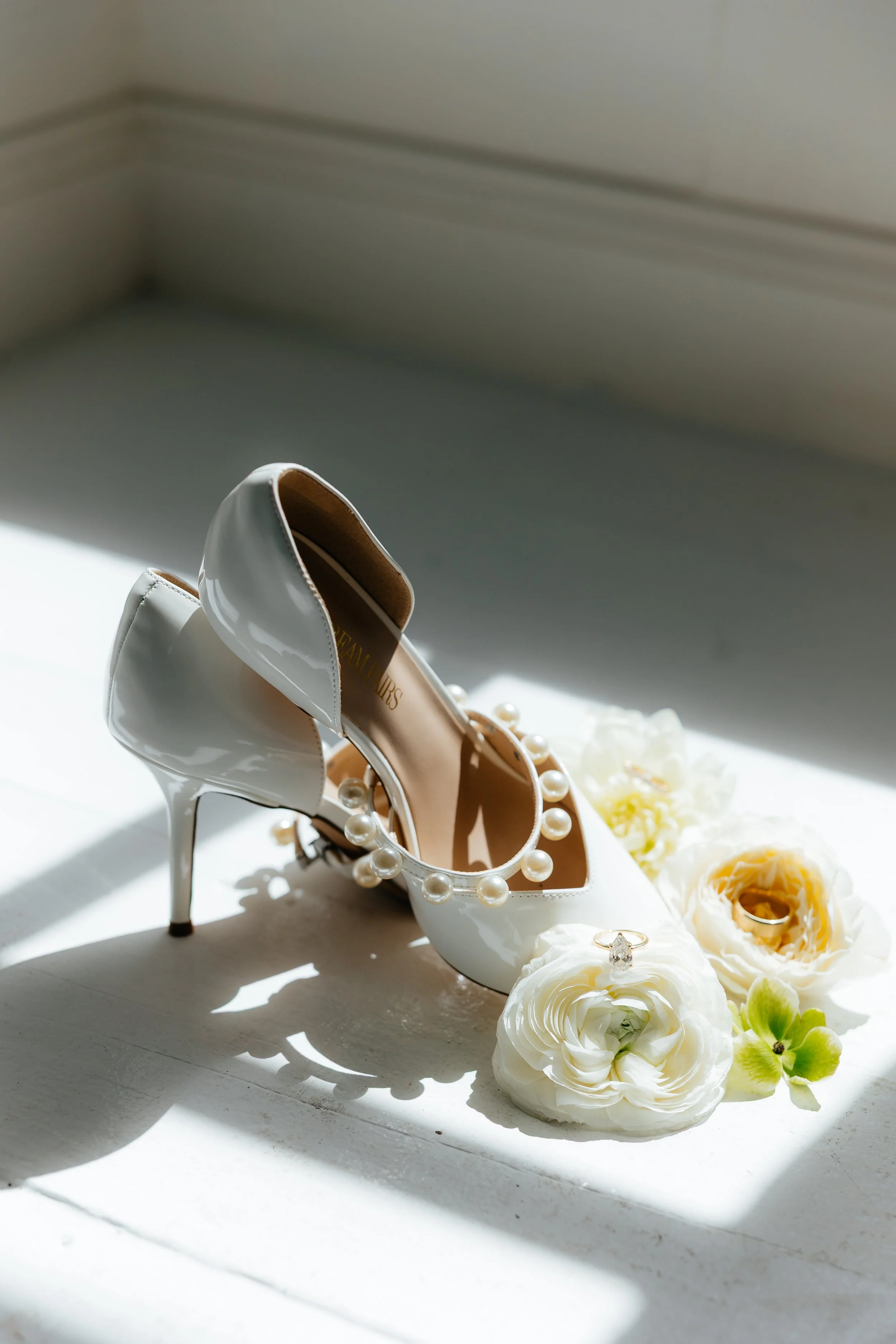 White high-heeled shoes with pearl embellishments, surrounded by white and green flowers, on a white surface with sunlight and shadows.