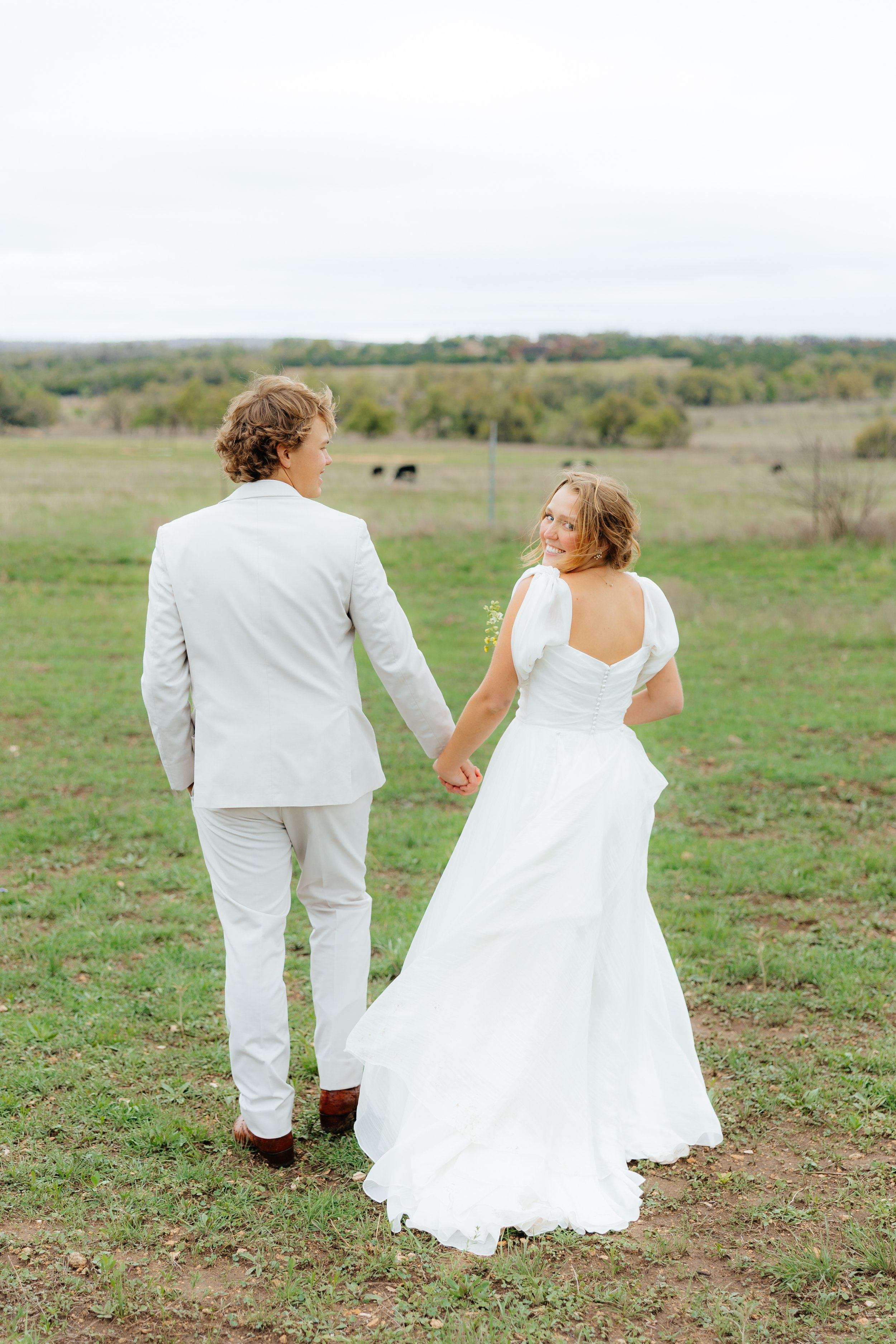 A young couple holding hands and walking in a field, with the woman smiling and looking back at the camera, dressed in a white wedding gown and the man in a white suit, during daytime.