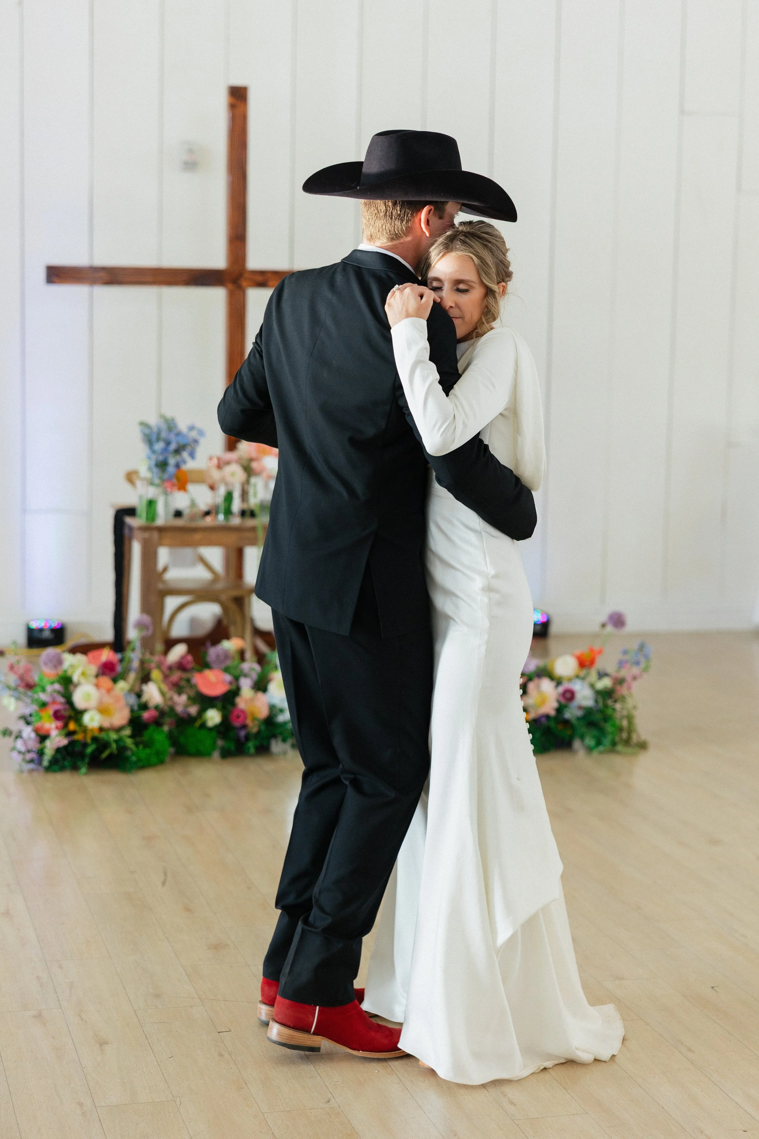 A couple shares a first dance at their wedding ceremony, with a cross and floral decorations in the background.