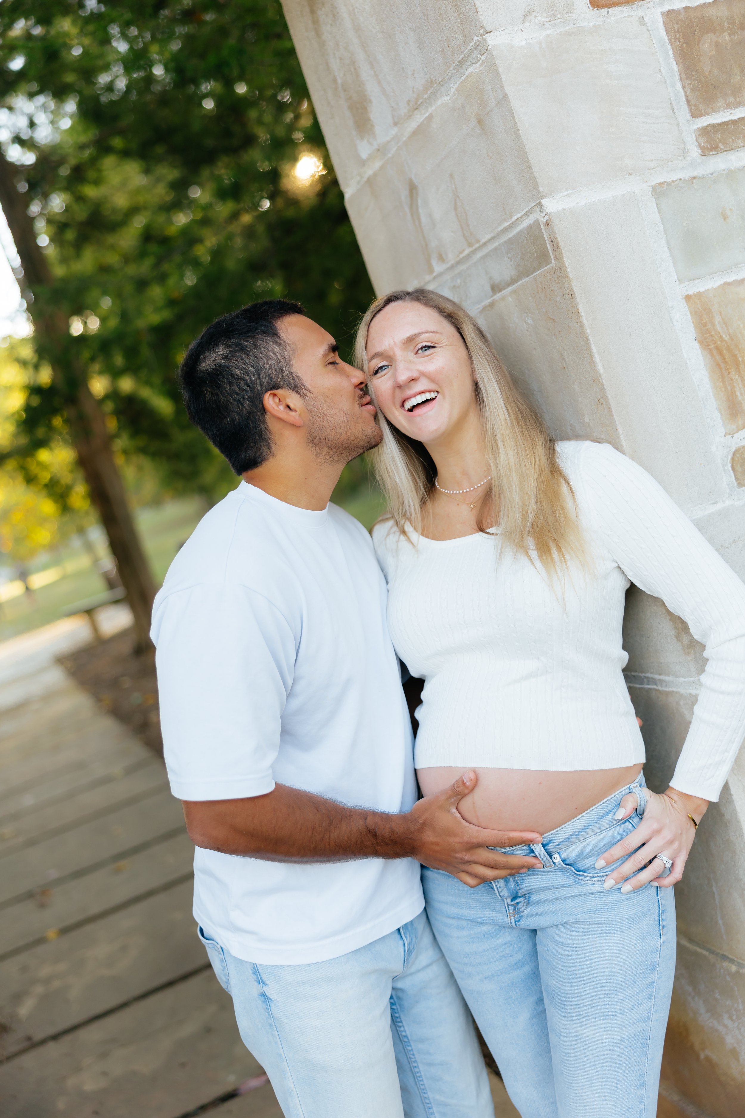 A couple standing outdoors, with the man whispering in the woman's ear as she smiles and touches her pregnant belly.