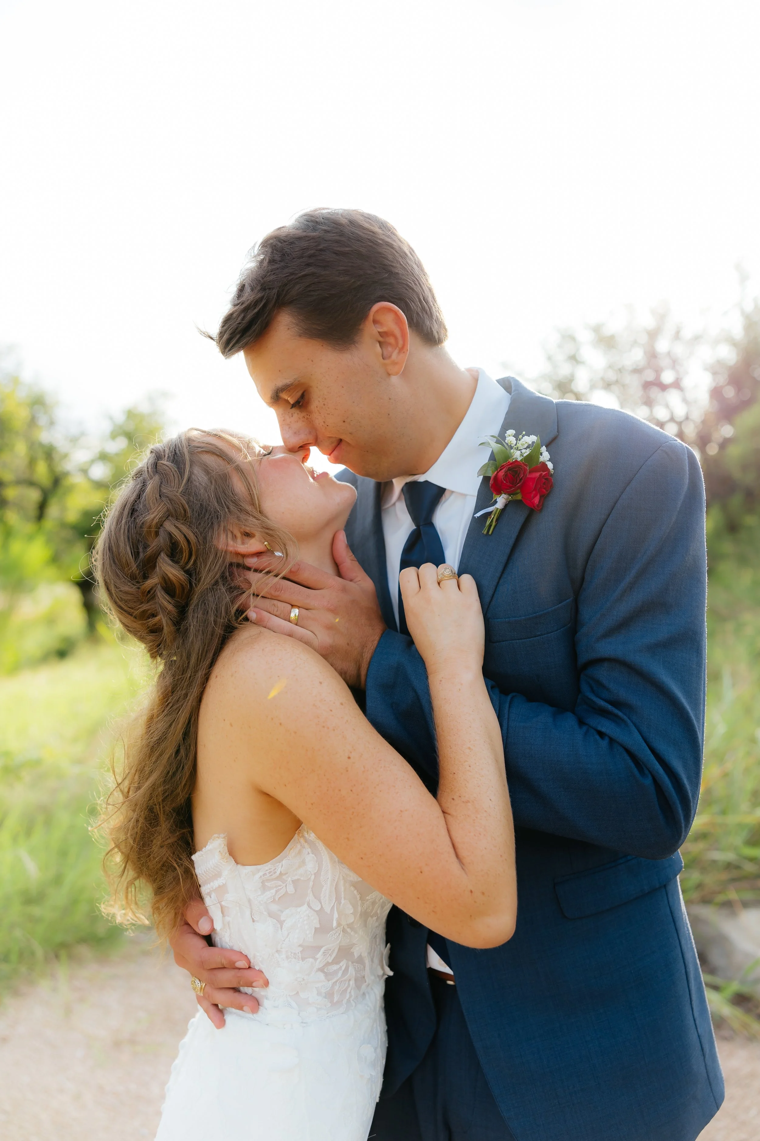 A bride and groom sharing a kiss outdoors on their wedding day, with the groom in a navy suit and the bride in a white lace dress.