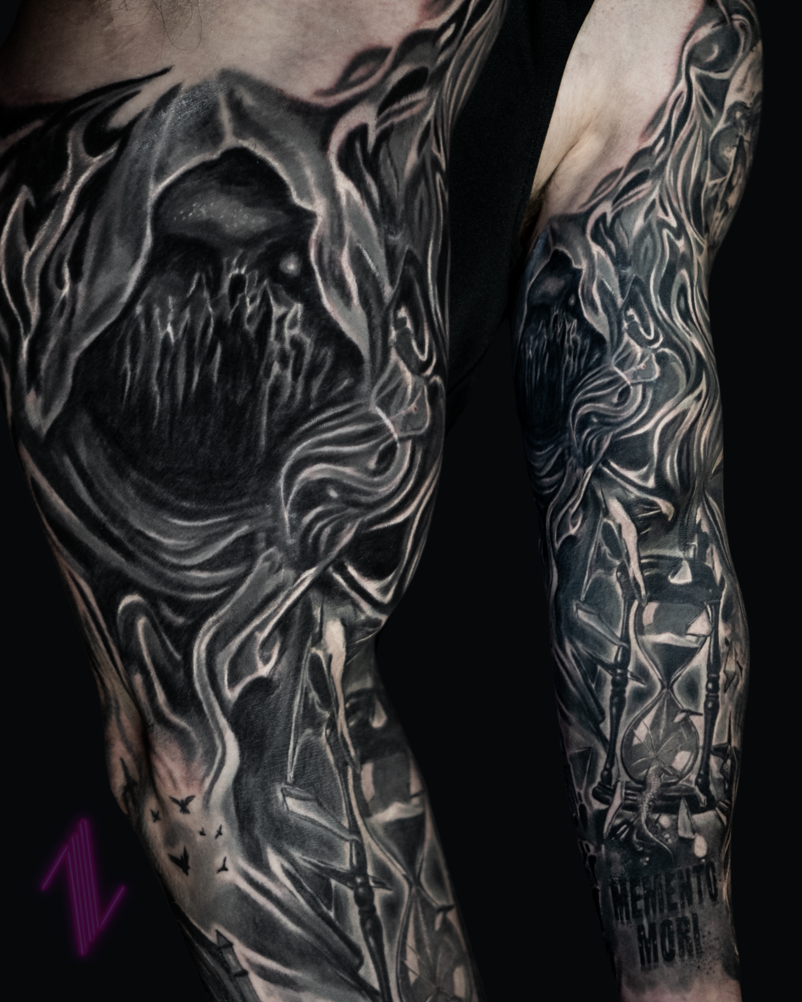 Close-up of black and grey tattoos on a person's arm and shoulder, featuring a stylized face with flowing lines, candles, and small bats.